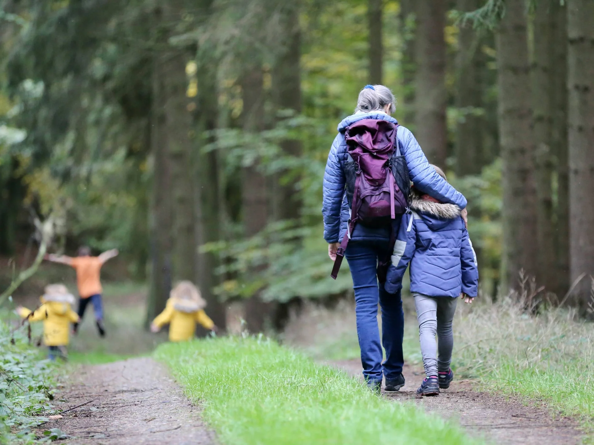 Frau mit Kind beim Spaziergang im Wald, andere Kinder spielen im Hintergrund