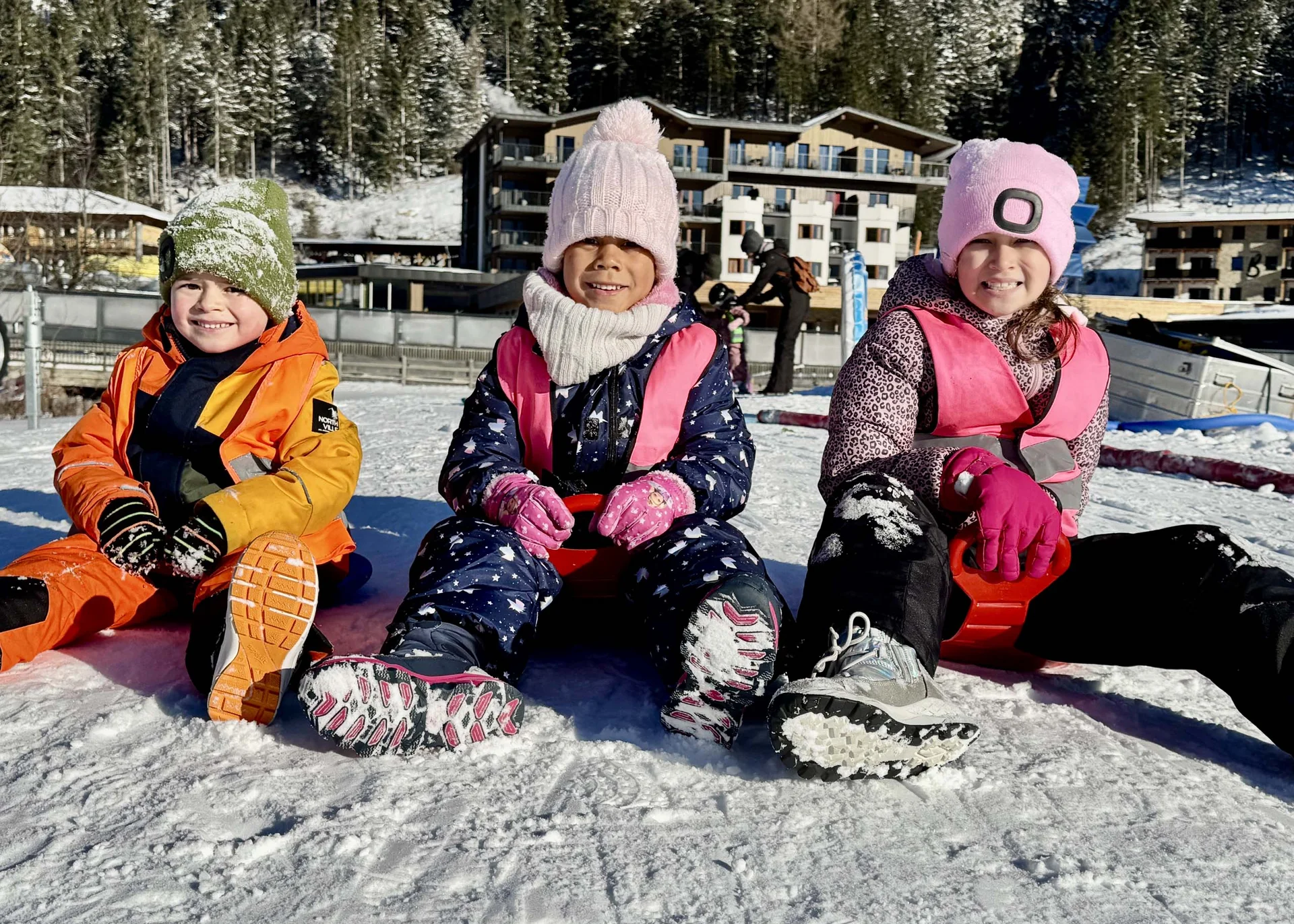 Drei Kinder in bunter Winterkleidung sitzen auf Schlitten im Schnee
