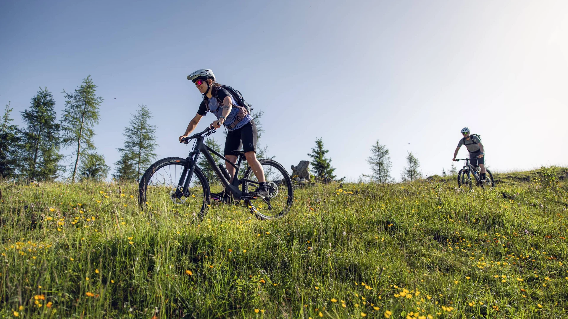 Zwei Frauen fahren Mountainbike auf einem blumenreichen Hügel bei sonnigem Wetter