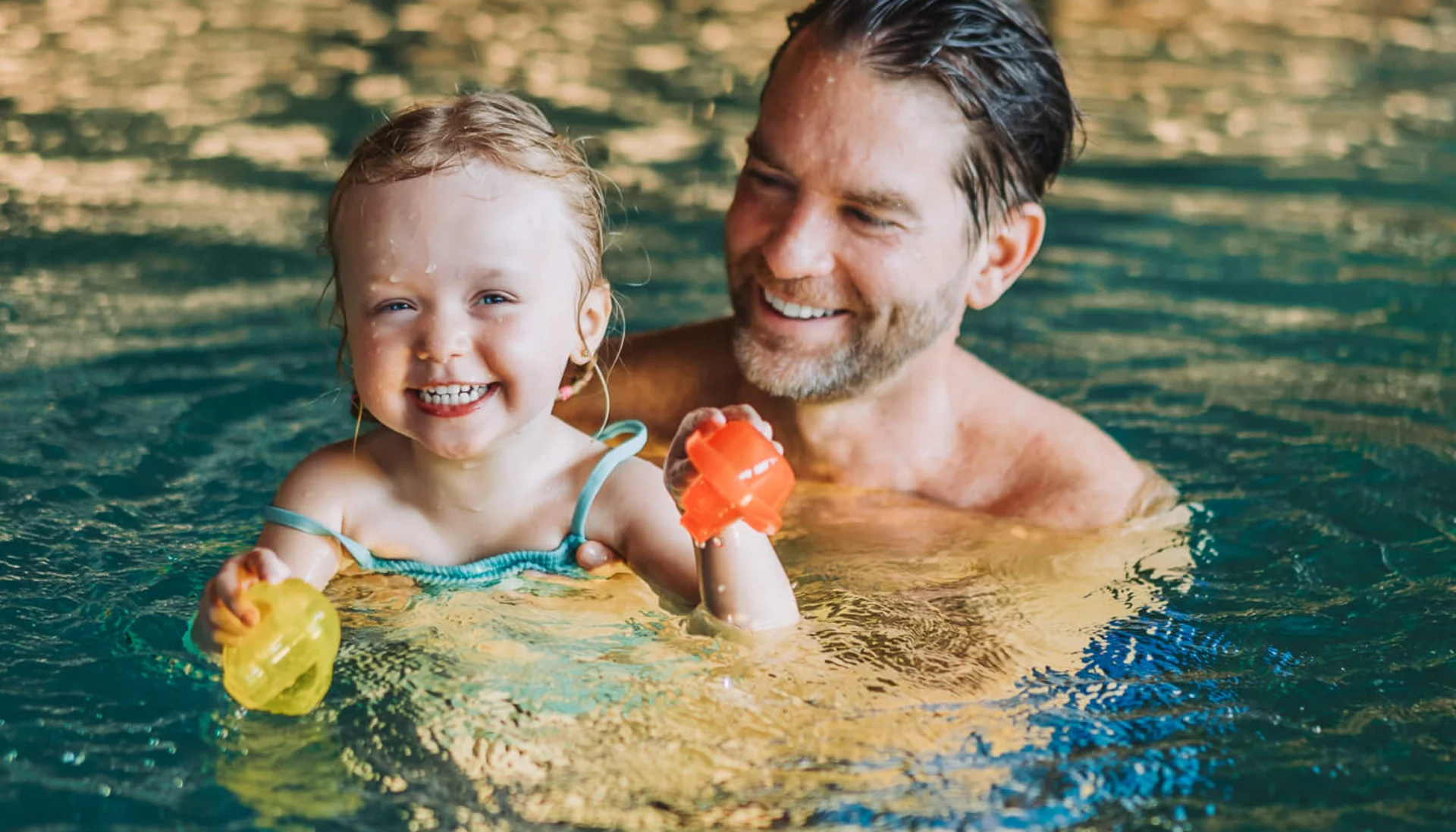 Vater und Tochter spielen lachend im Schwimmbecken mit Wasserspielzeug