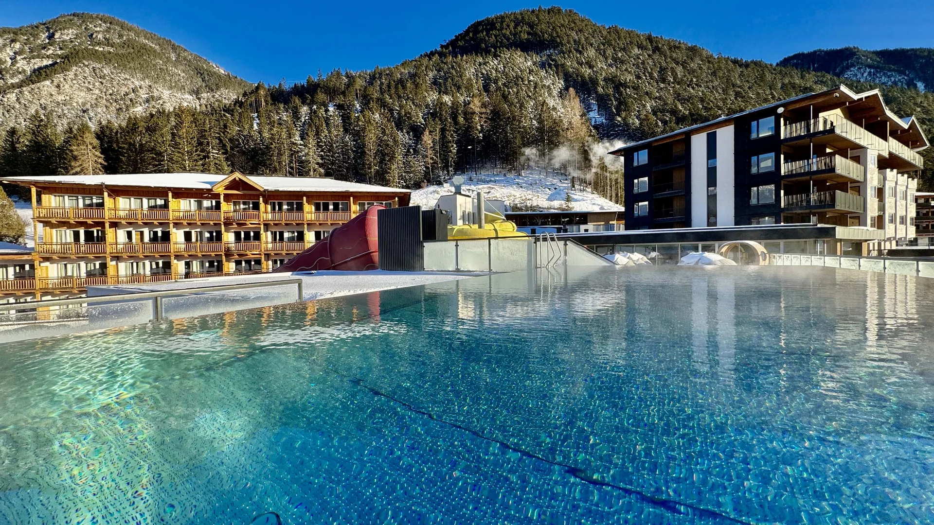 Beheizter Außenpool mit Bergkulisse und Gebäuden im Hintergrund unter klarem Himmel