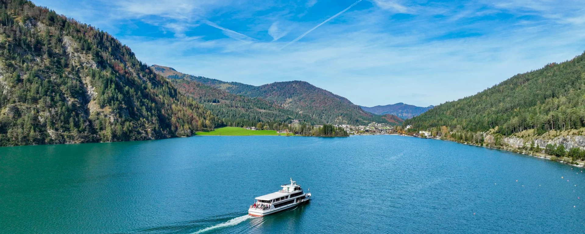 Fähre auf blauem See umgeben von bewaldeten Bergen unter blauem Himmel