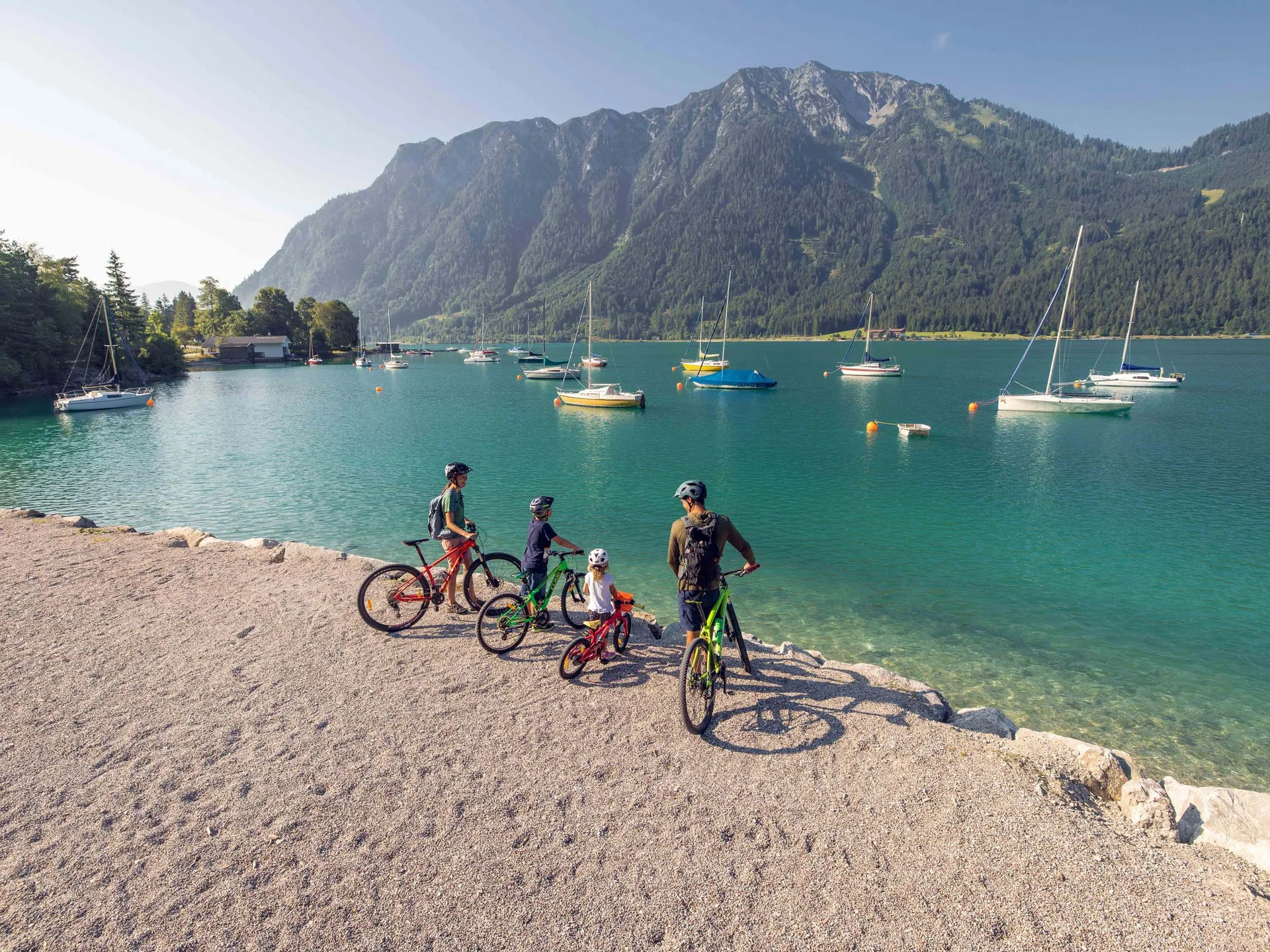 Familie mit Fahrrädern am Seeufer vor Bergen und segelbooten