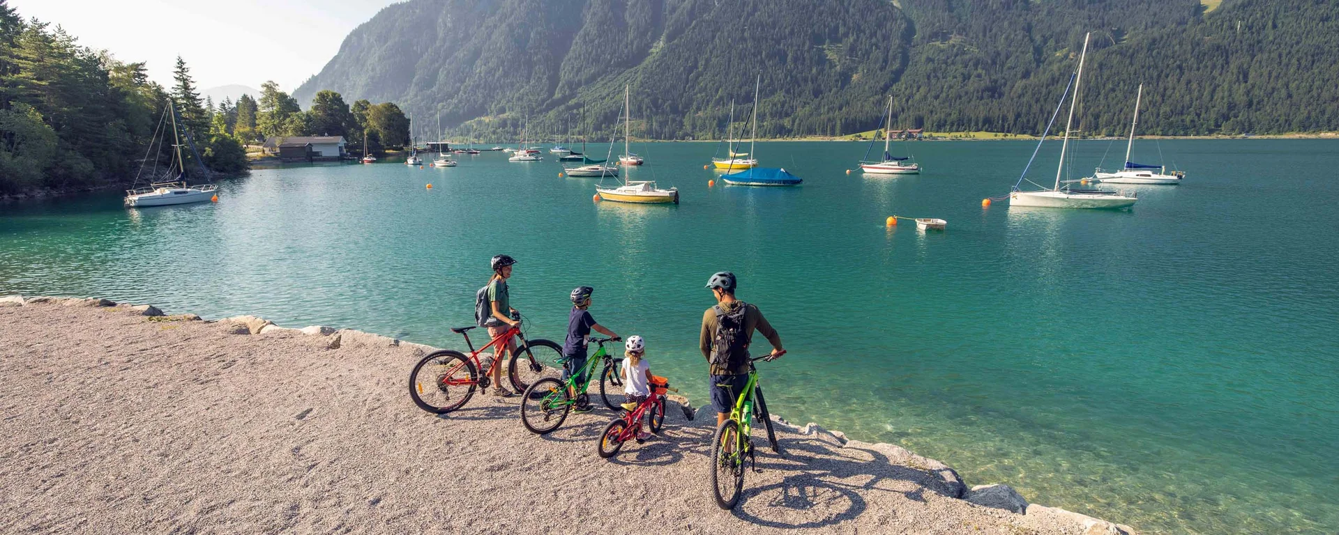 Familie mit Fahrrädern am Seeufer vor Bergen und segelbooten