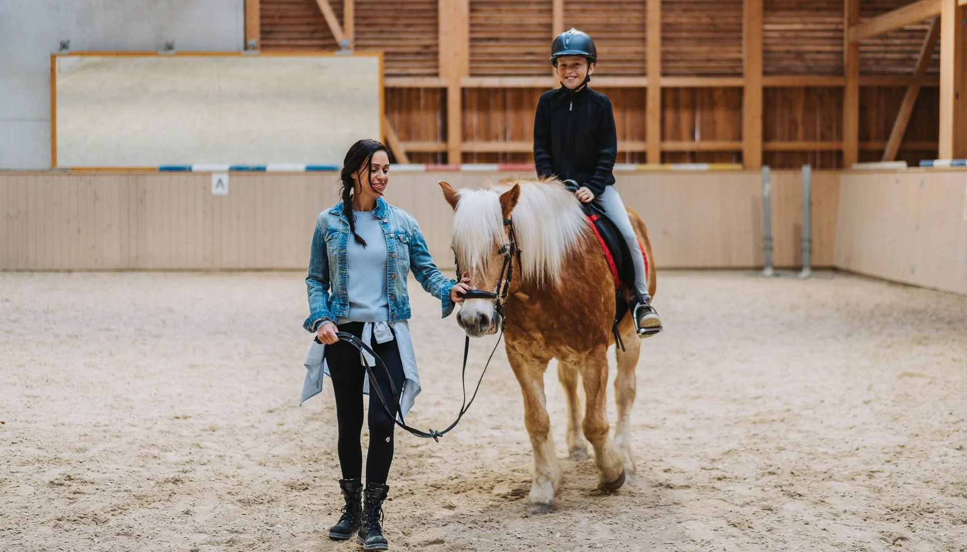 Frau führt Pony, Kind reitet mit Helm in Reithalle