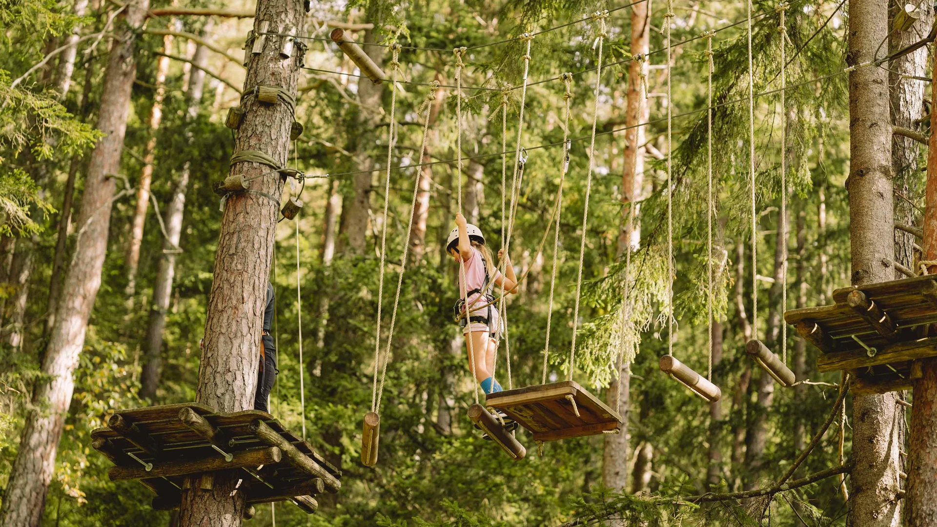 Kind mit Helm klettert im Hochseilgarten zwischen Bäumen