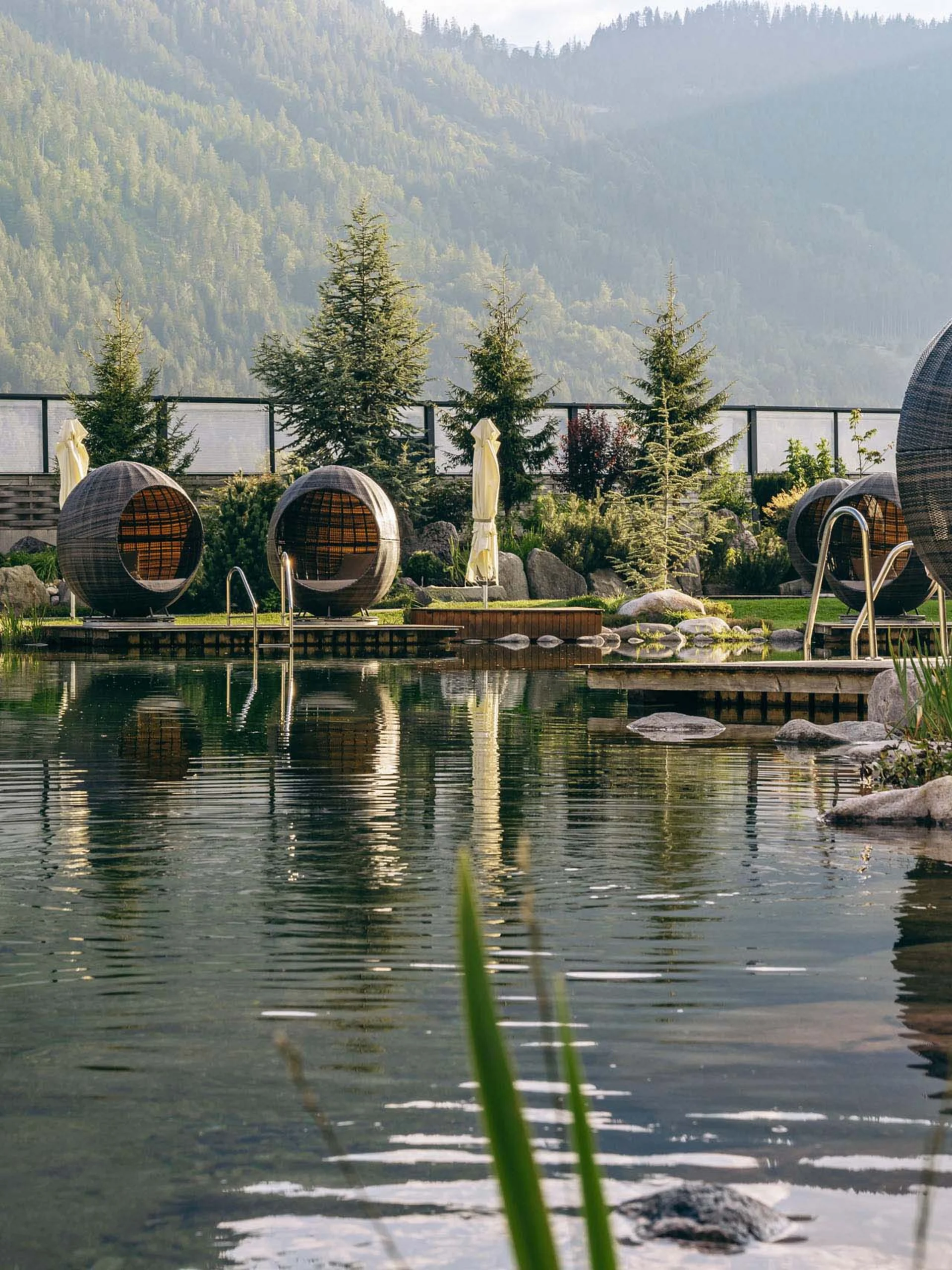 Natürlicher Pool mit runden Sitzpods in einer Gartenanlage mit Bergen im Hintergrund