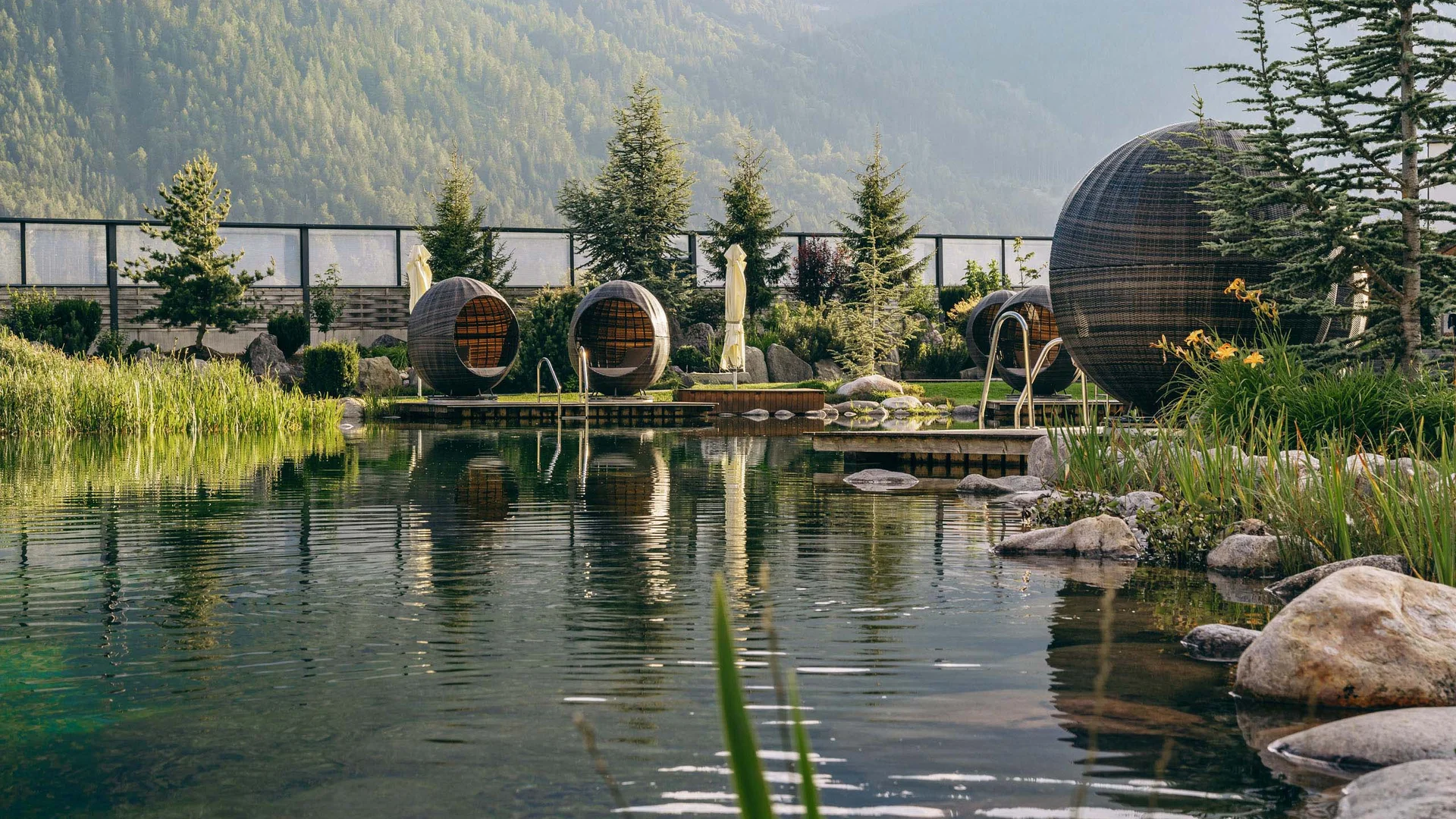 Natürlicher Pool mit runden Sitzpods in einer Gartenanlage mit Bergen im Hintergrund