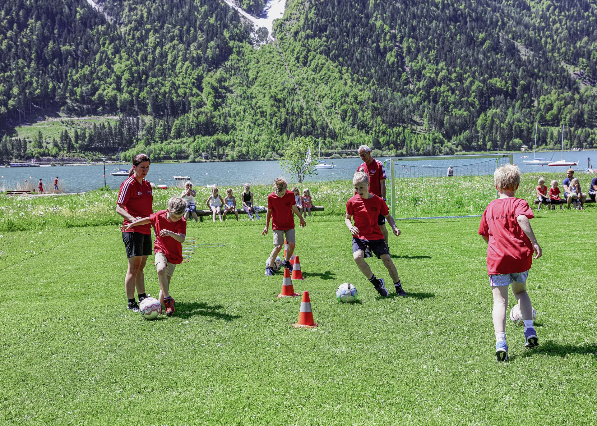 Kinder trainieren Fußball mit Trainern auf Wiese mit Bergen und See im Hintergrund