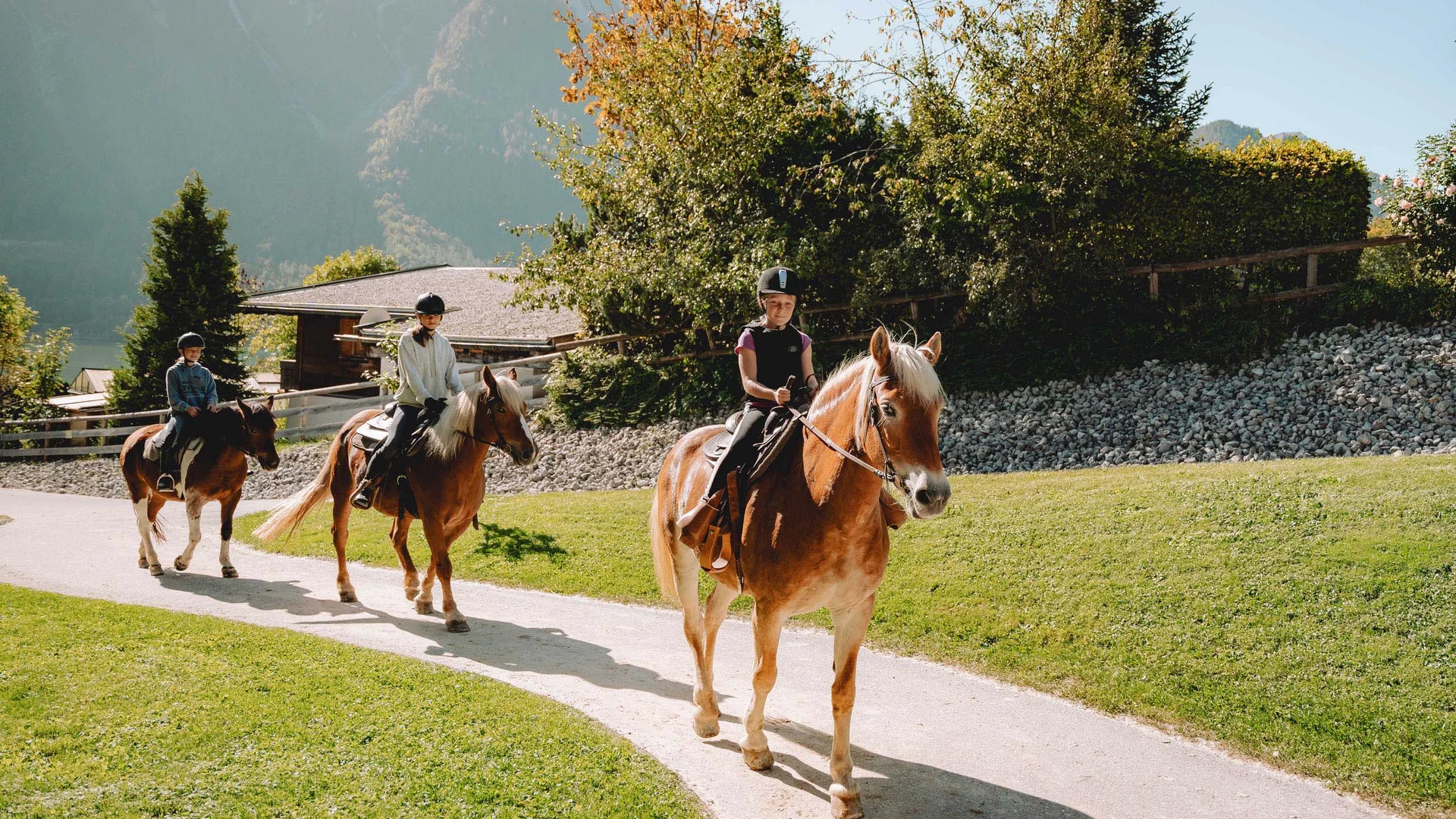 Drei Kinder reiten auf Pferden auf einem Weg vor einer Berglandschaft