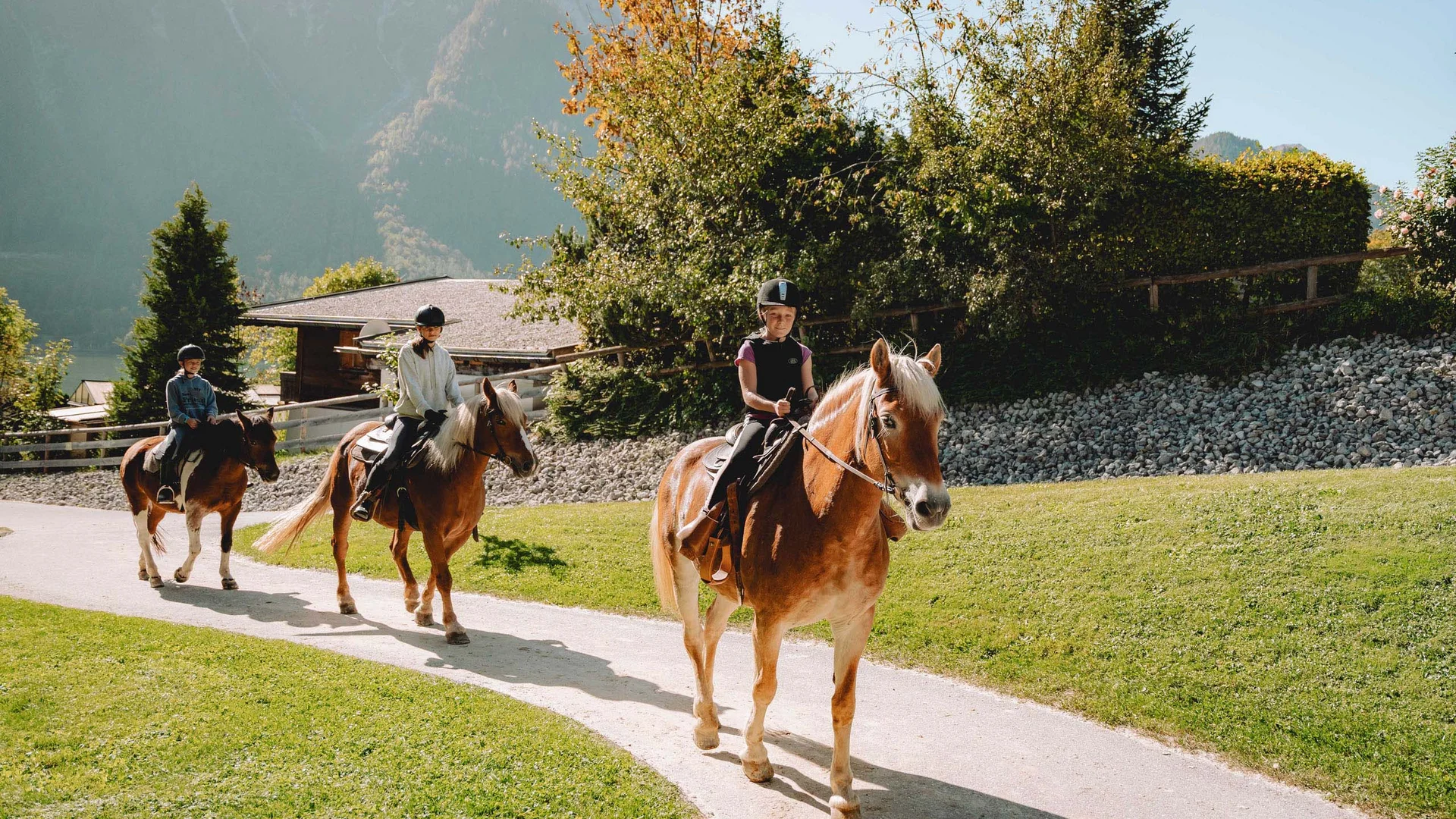 Drei Kinder reiten auf Pferden auf einem Weg vor einer Berglandschaft