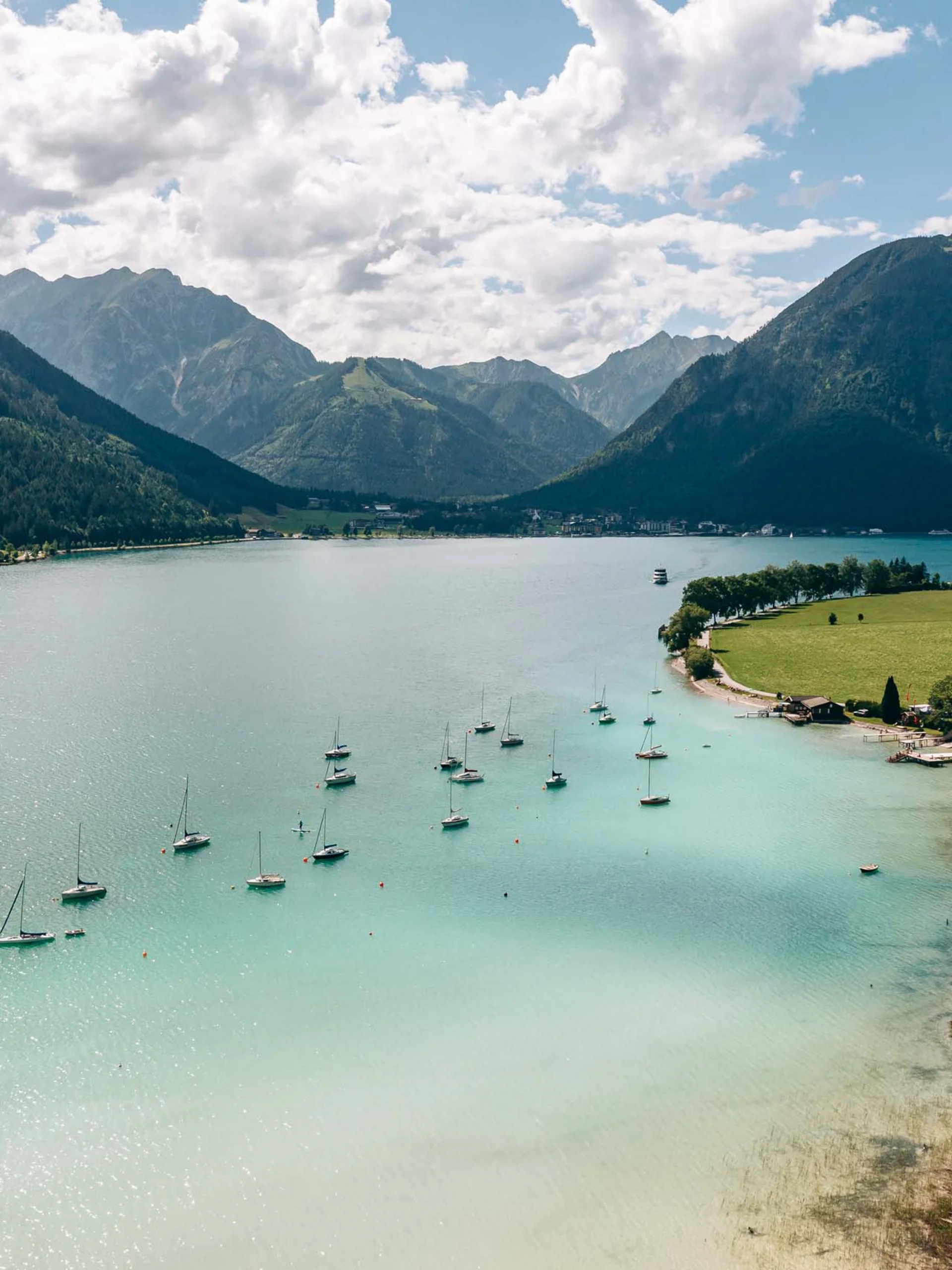 Blick auf einen See mit Segelbooten und Bergen im Hintergrund bei sonnigem Himmel