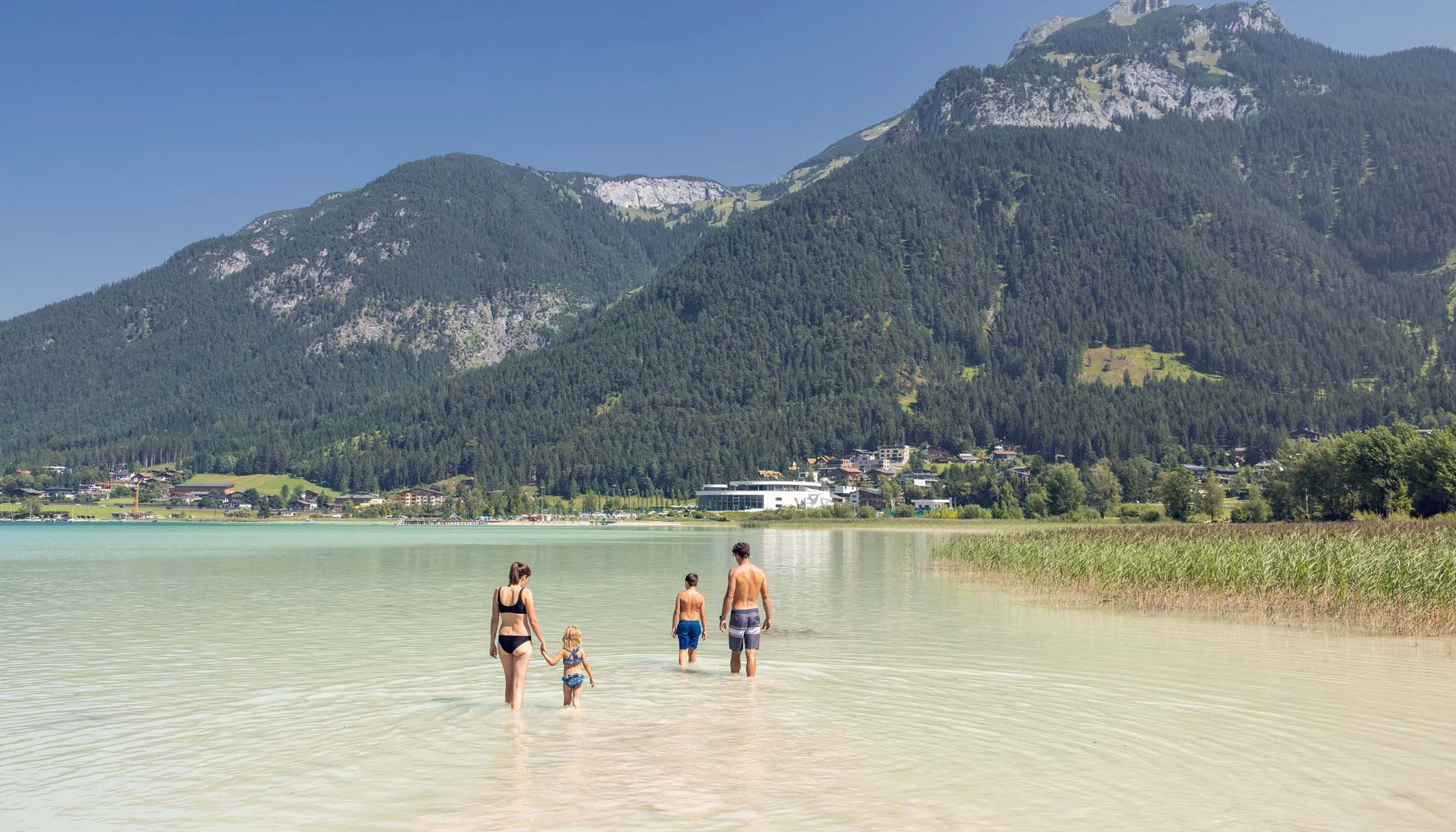 Familie watet in klarem Wasser vor Bergkulisse und Dorf im Hintergrund