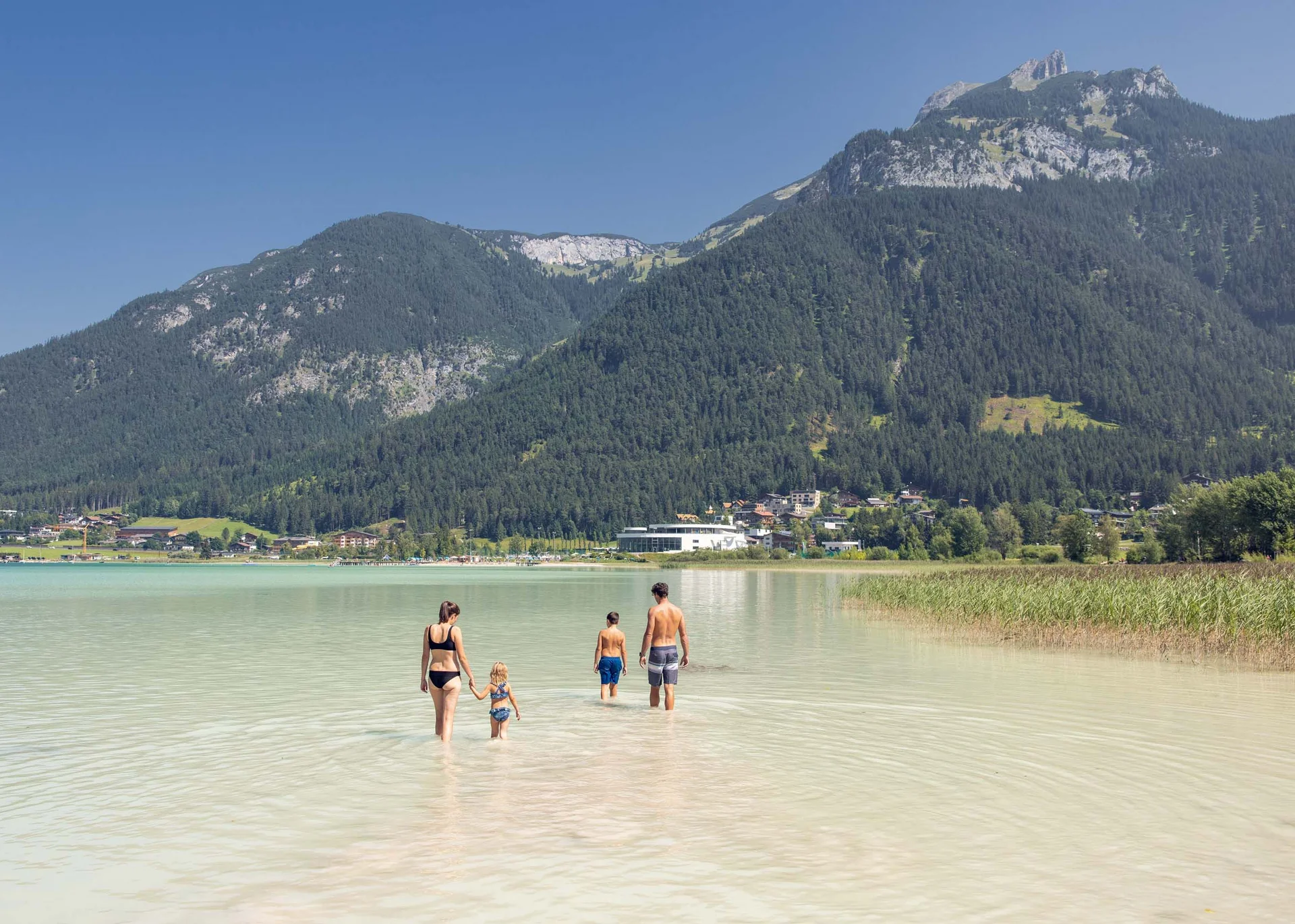 Familie watet in klarem Wasser vor Bergkulisse und Dorf im Hintergrund