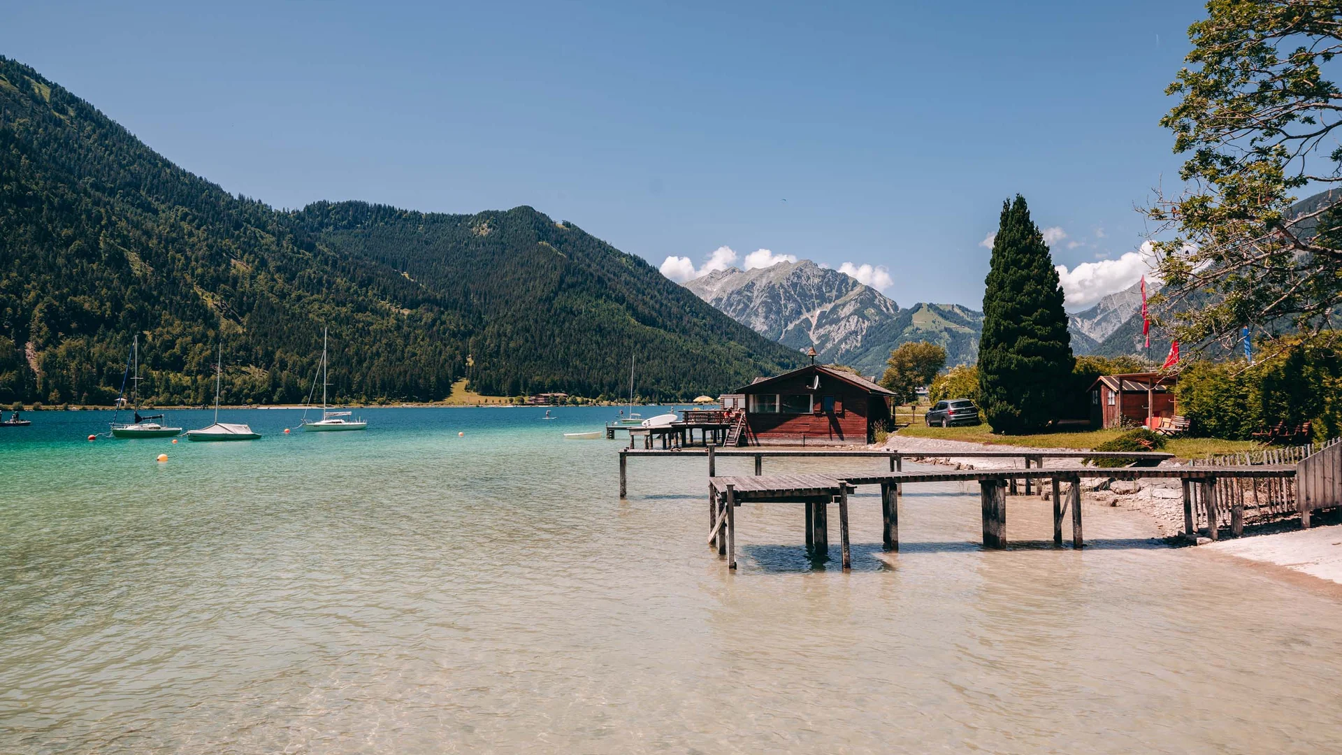 Klares Wasser, Boote und Holzanleger an einem Bergsee mit Hütten und Bergen im Hintergrund