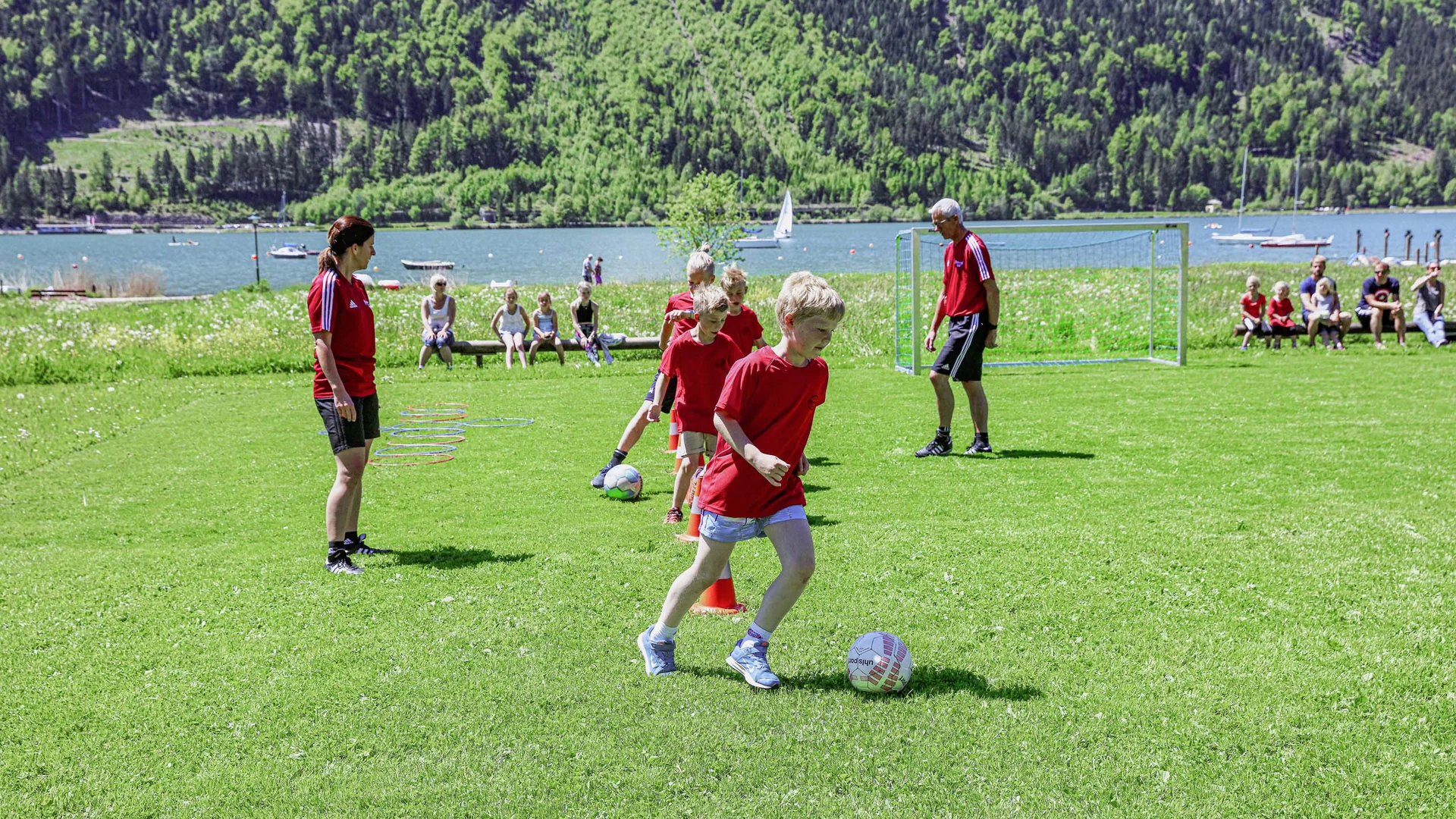 Kinder trainieren Fußball mit Trainern auf einer Wiese am See vor Zuschauern