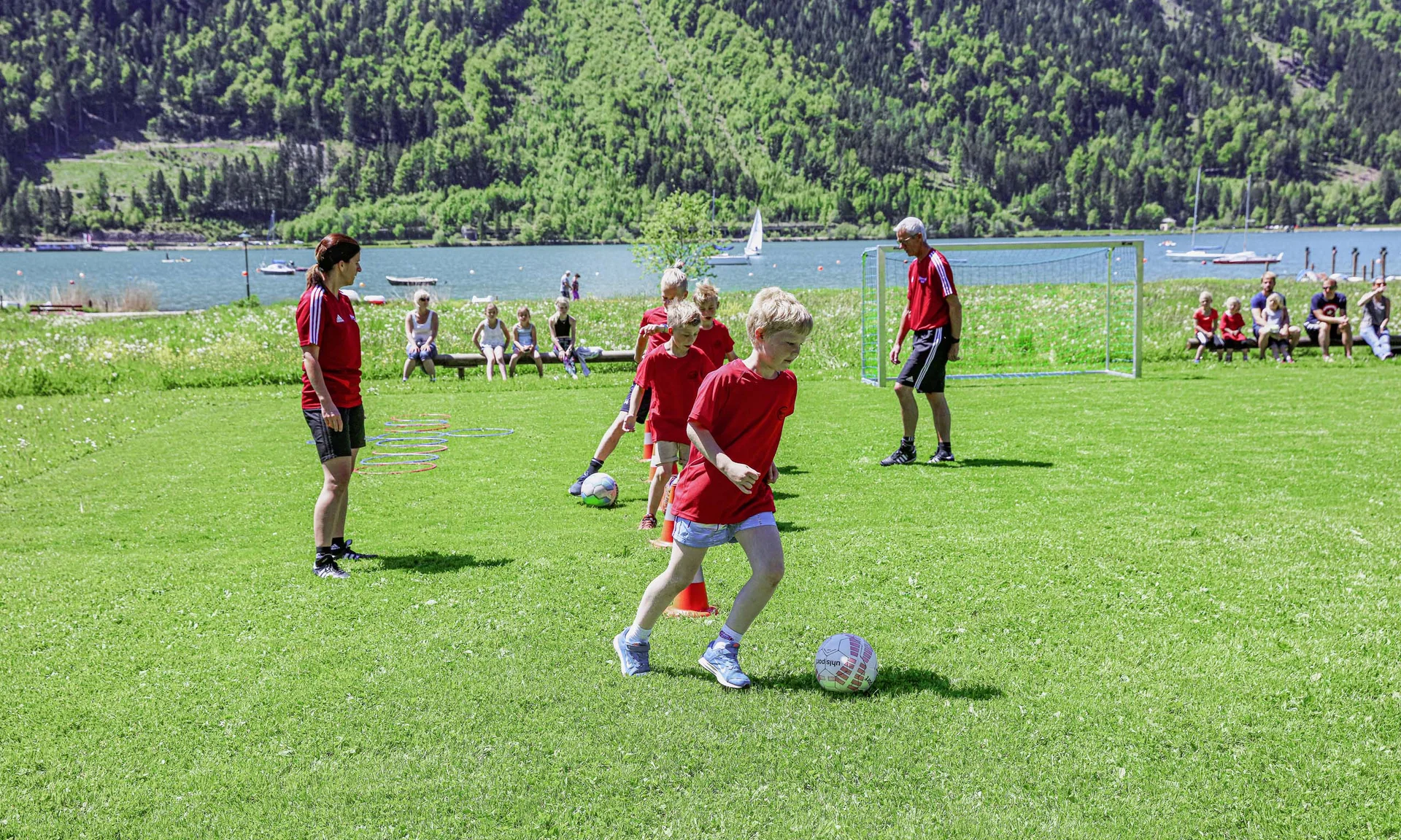 Kinder trainieren Fußball mit Trainern auf einer Wiese am See vor Zuschauern