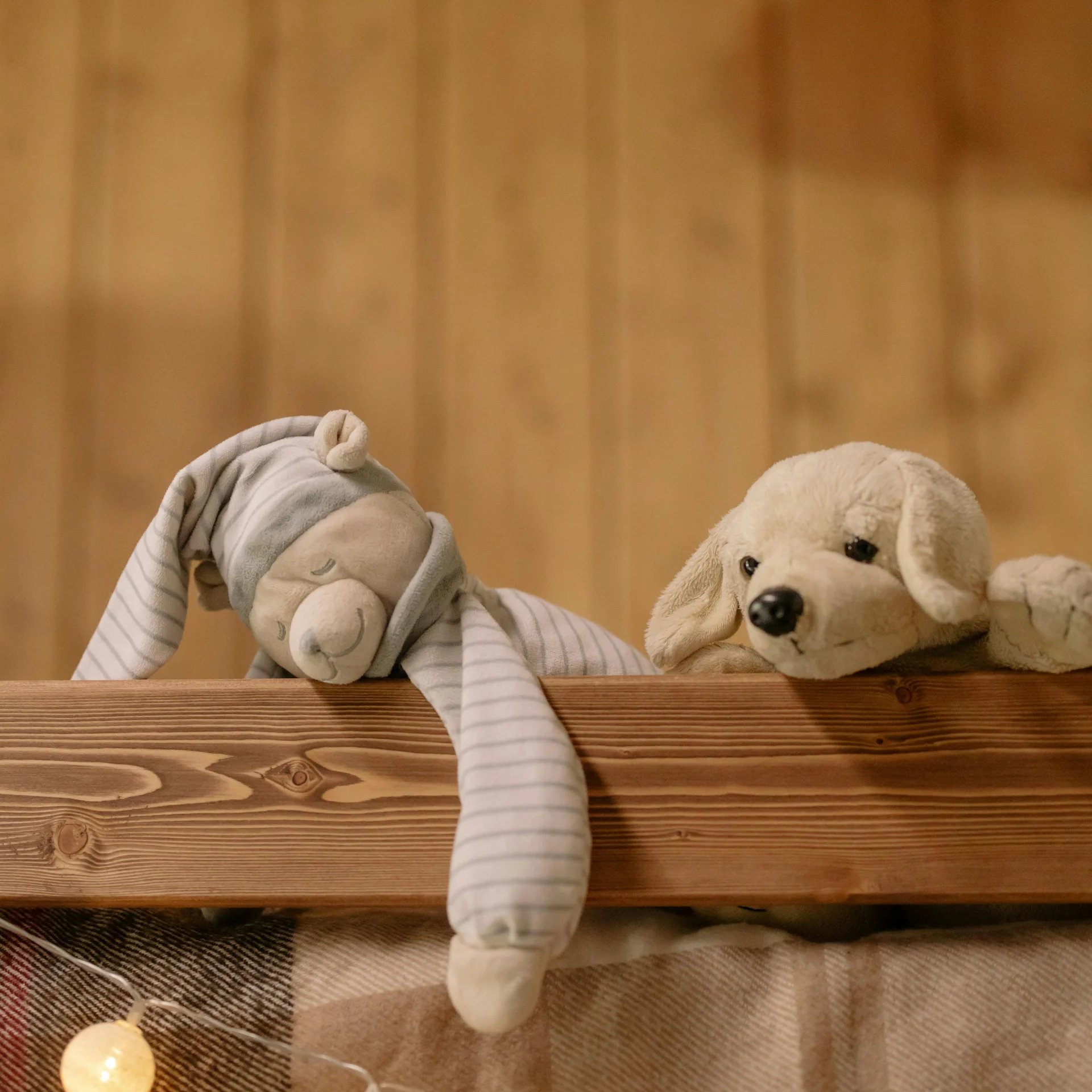 Two stuffed animals on a wooden beam above a plaid blanket with string lights