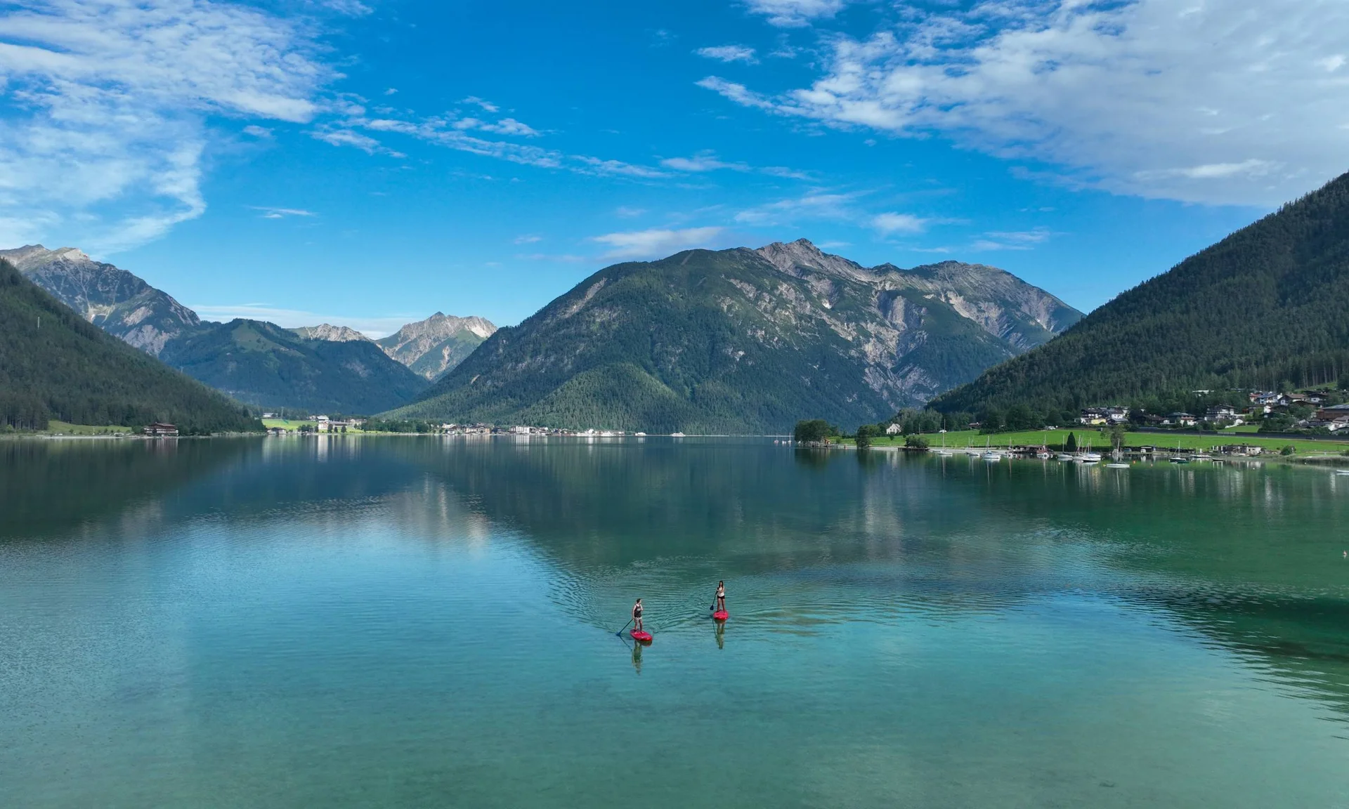 Zwei Personen paddeln auf einem See mit Bergen und Dorf im Hintergrund