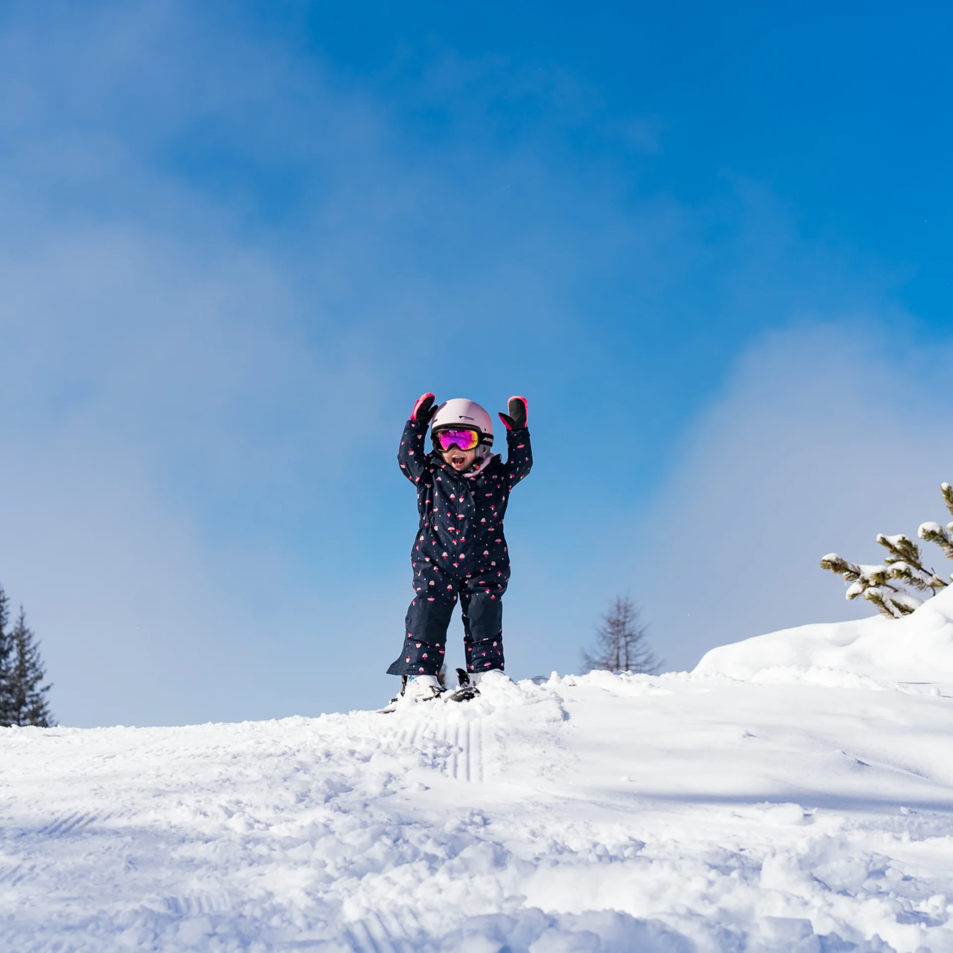 Kind in Skikleidung hebt die Arme auf schneebedecktem Hügel bei blauem Himmel