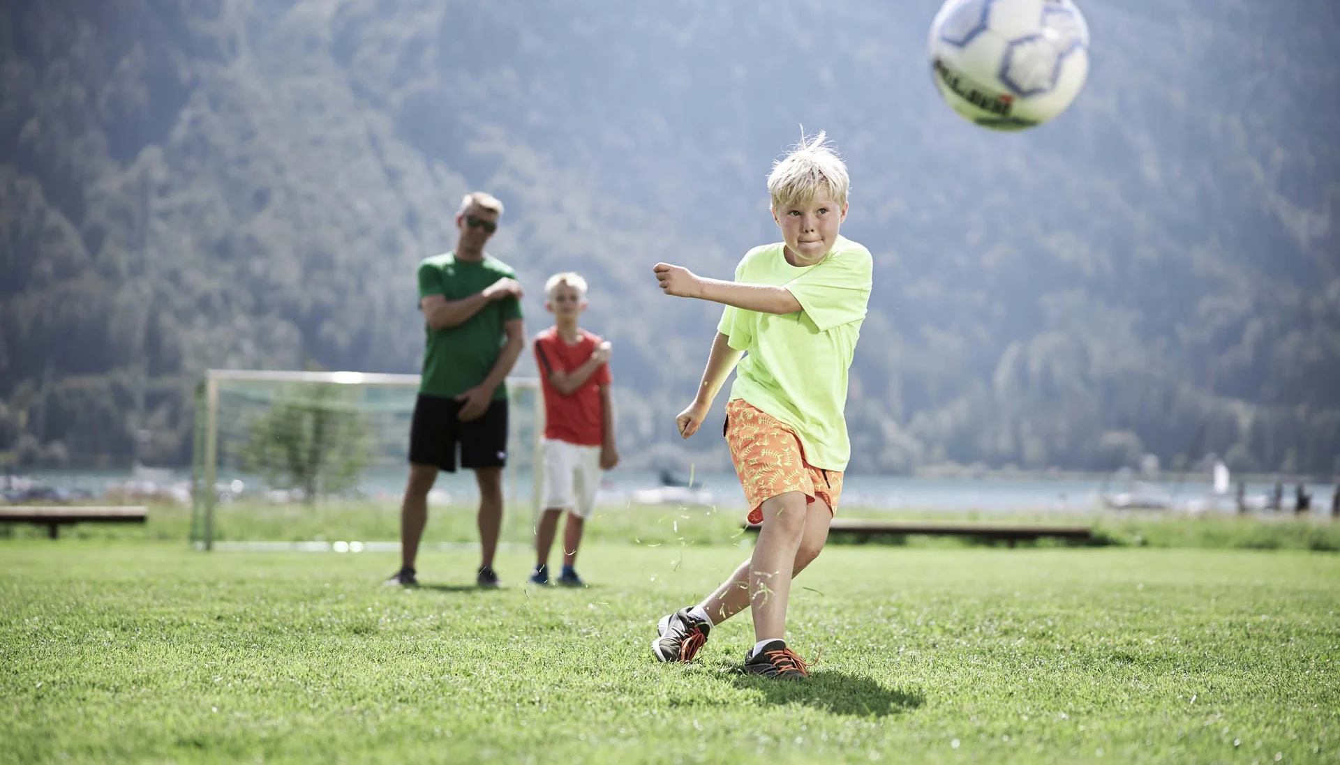 Junge schießt Fußball auf grünem Feld mit zwei Personen im Hintergrund