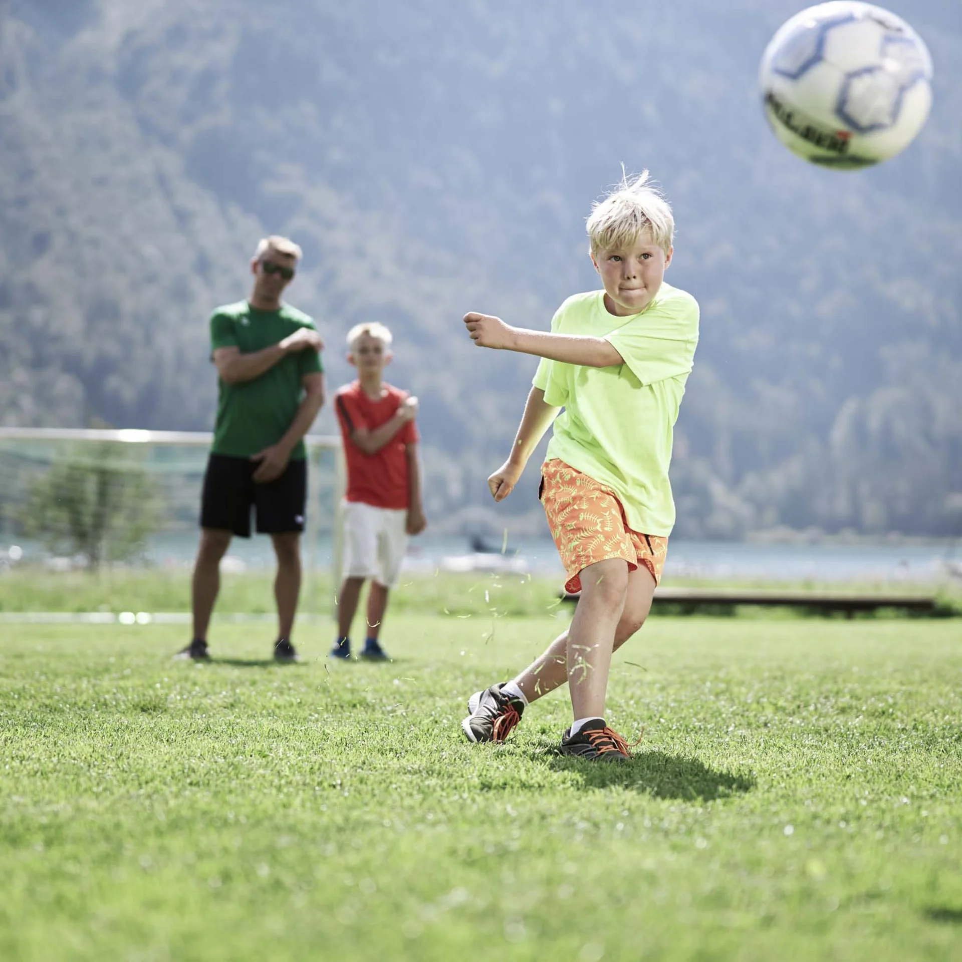Junge schießt Fußball auf grünem Feld mit zwei Personen im Hintergrund