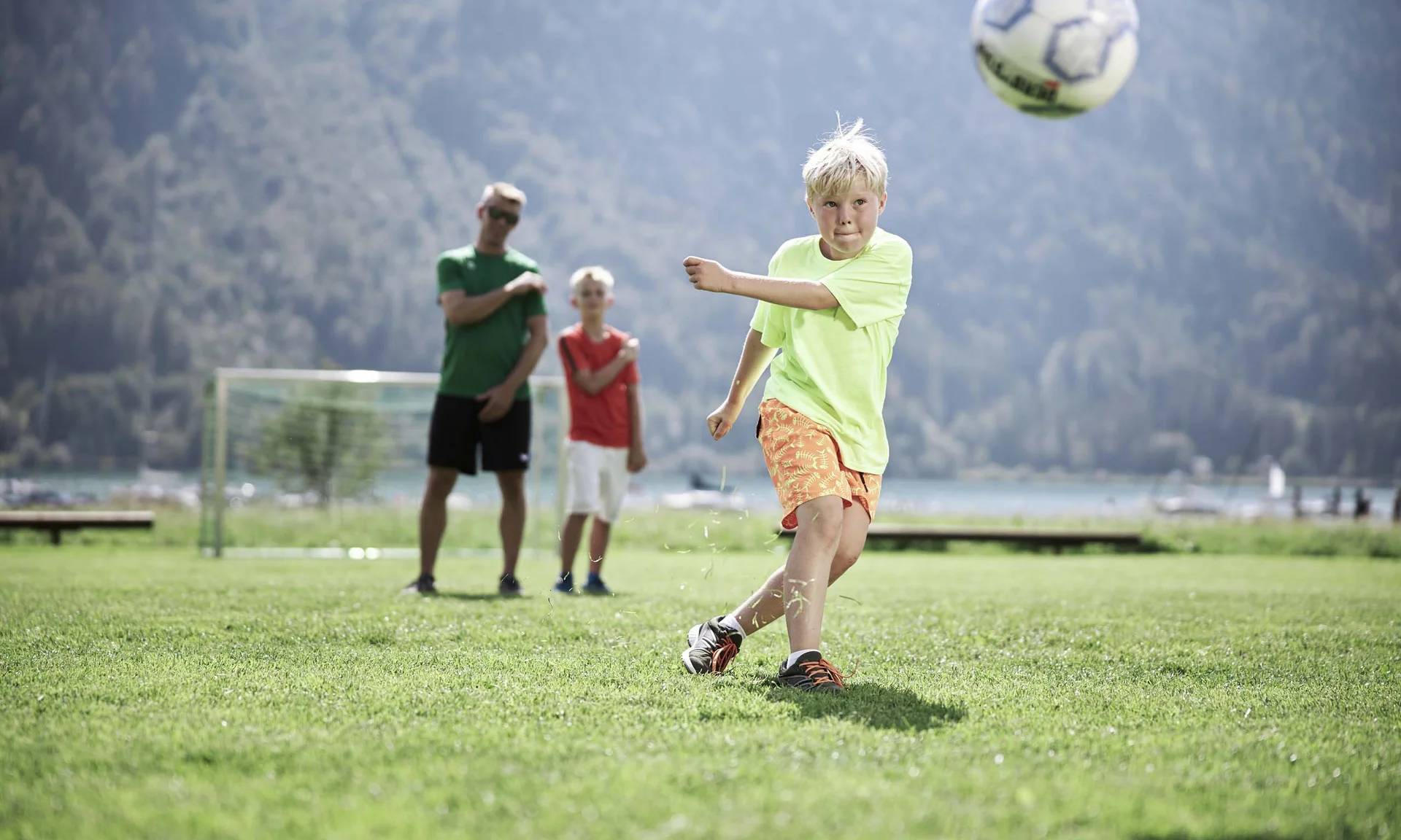 Junge schießt Fußball auf grünem Feld mit zwei Personen im Hintergrund