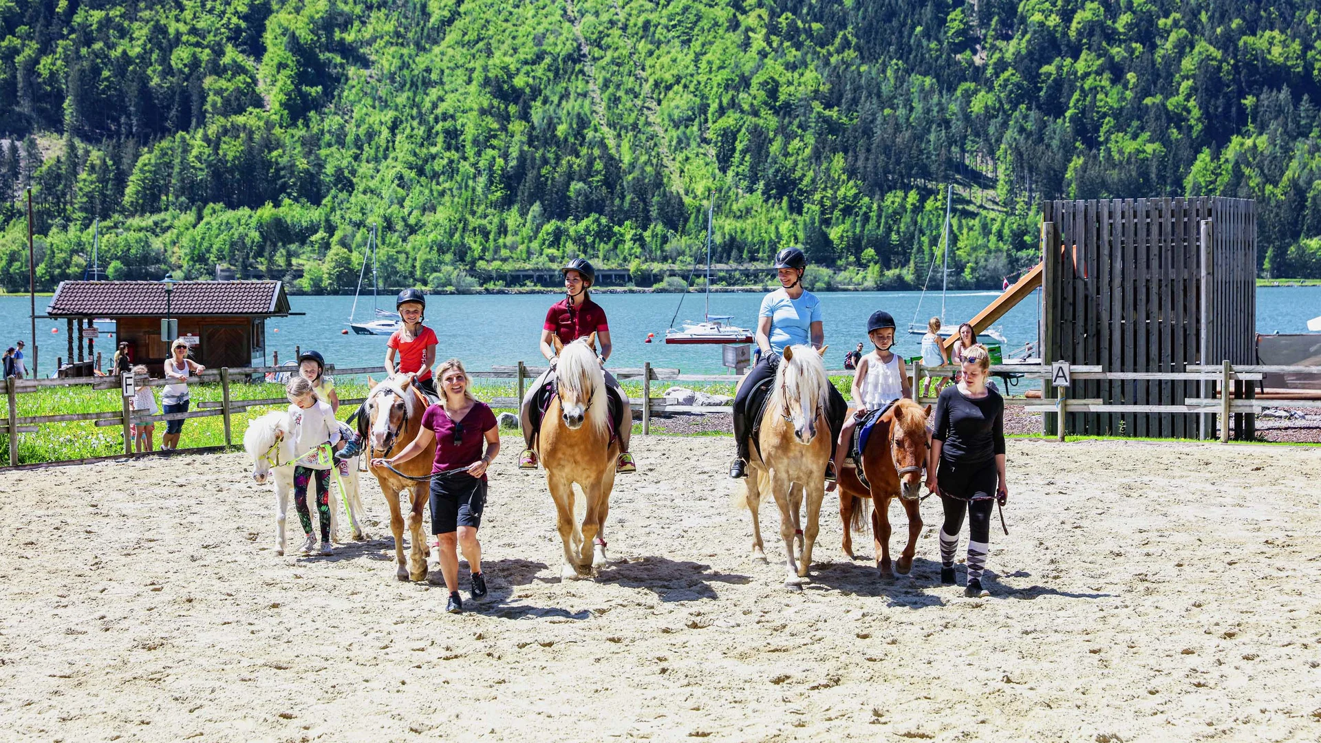 Kinder reiten Pferde in einer sonnigen Arena vor einem See und bewaldeten Hügeln