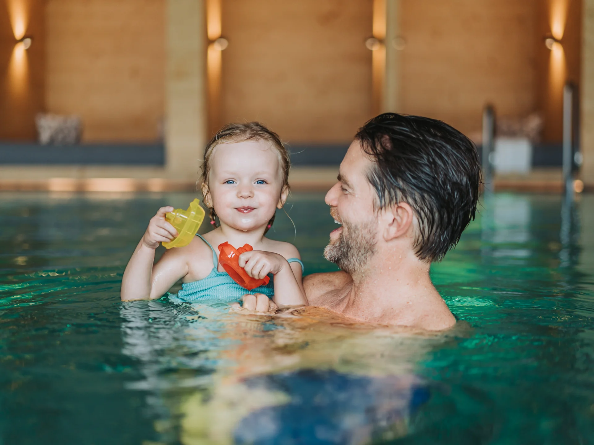 Vater hält lachendes kleines Mädchen mit Spielzeugbooten im Hallenbad