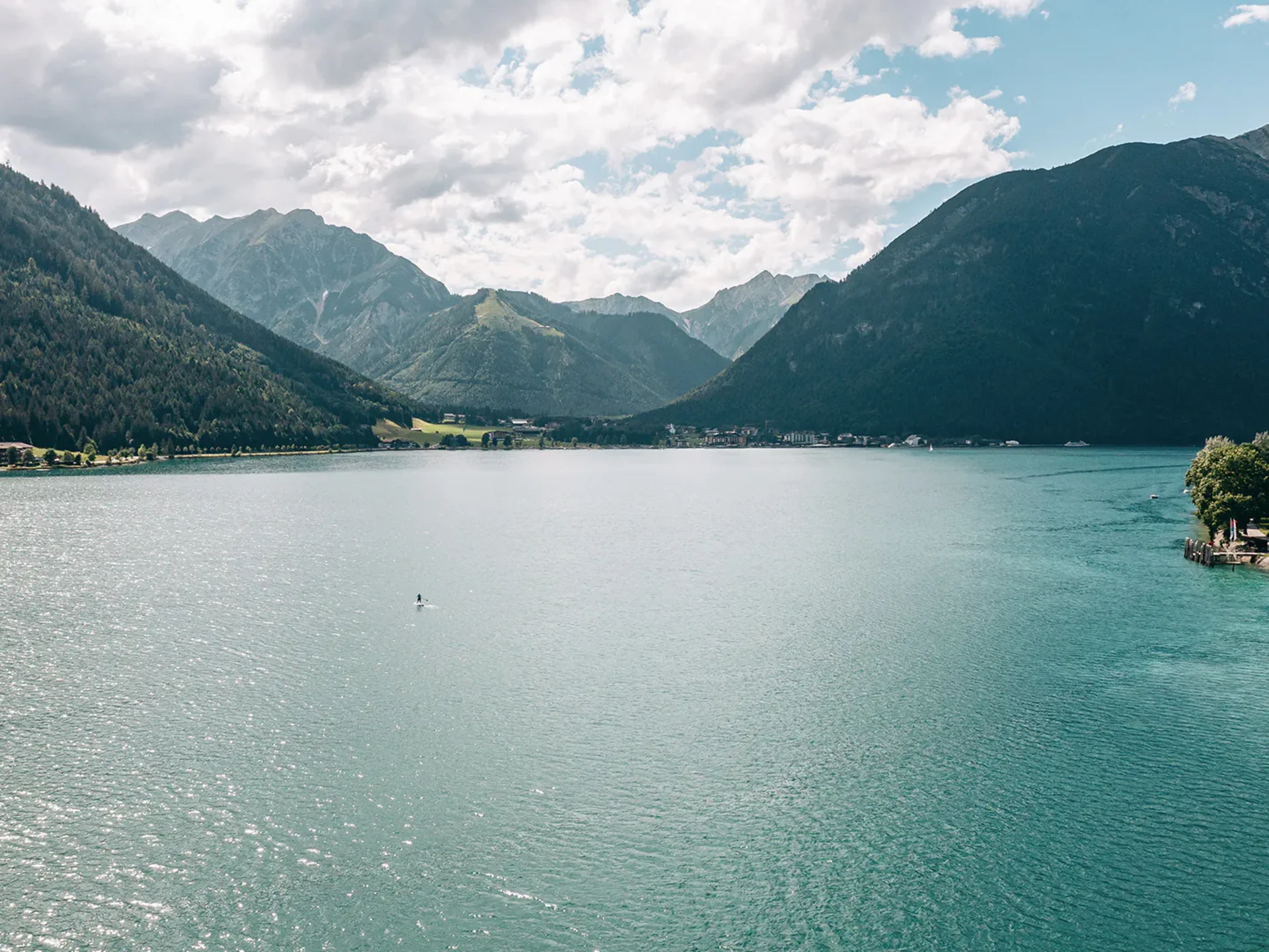 Bergsee mit bewaldeten Bergen, leicht bewölktem Himmel und einer Person auf einem Stand-up-Paddleboard