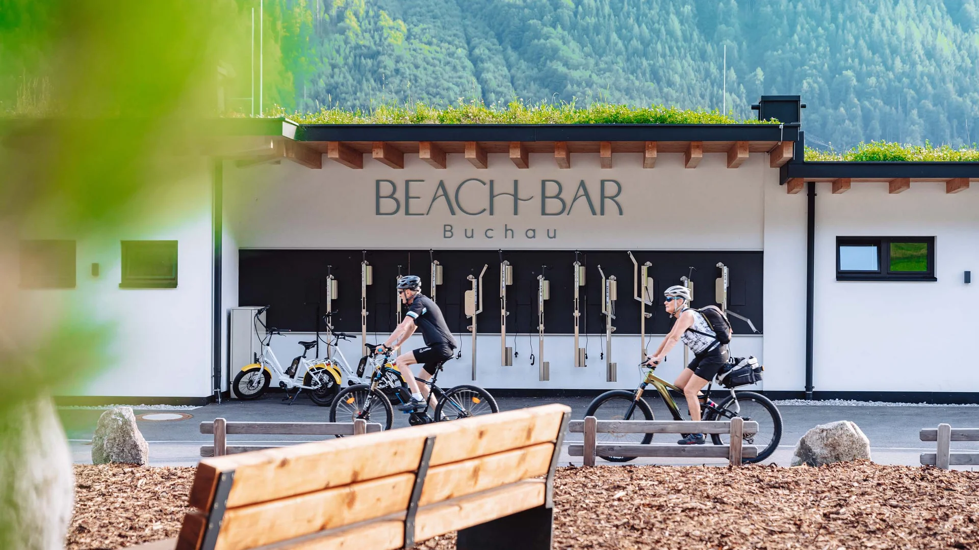 Zwei Radfahrer vor der Beach Bar Buchau mit grünen Bergen im Hintergrund