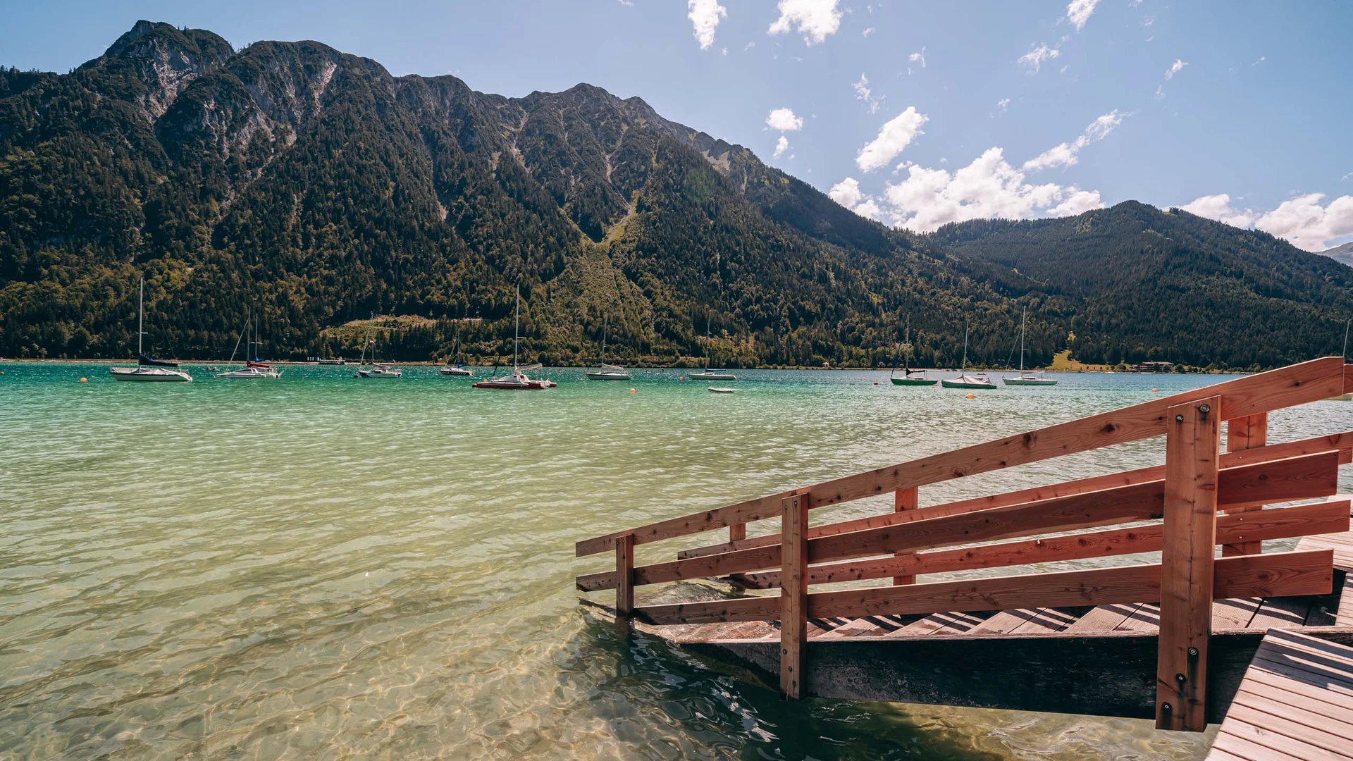 Bergsee mit Holzsteg und Booten vor bewaldeten Bergen bei klarem Himmel