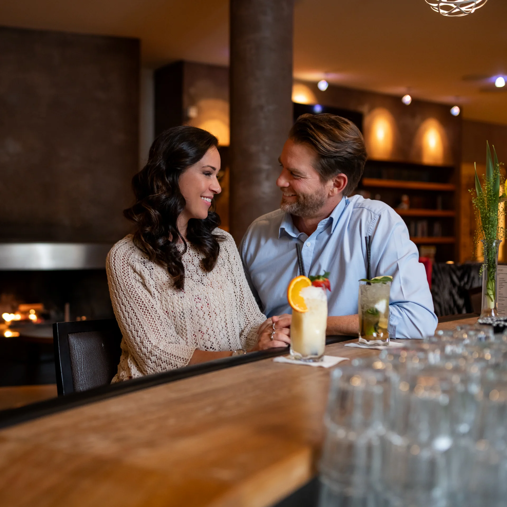 Paar sitzt an Bar mit Cocktails in gemütlicher Bar mit Kamin im Hintergrund