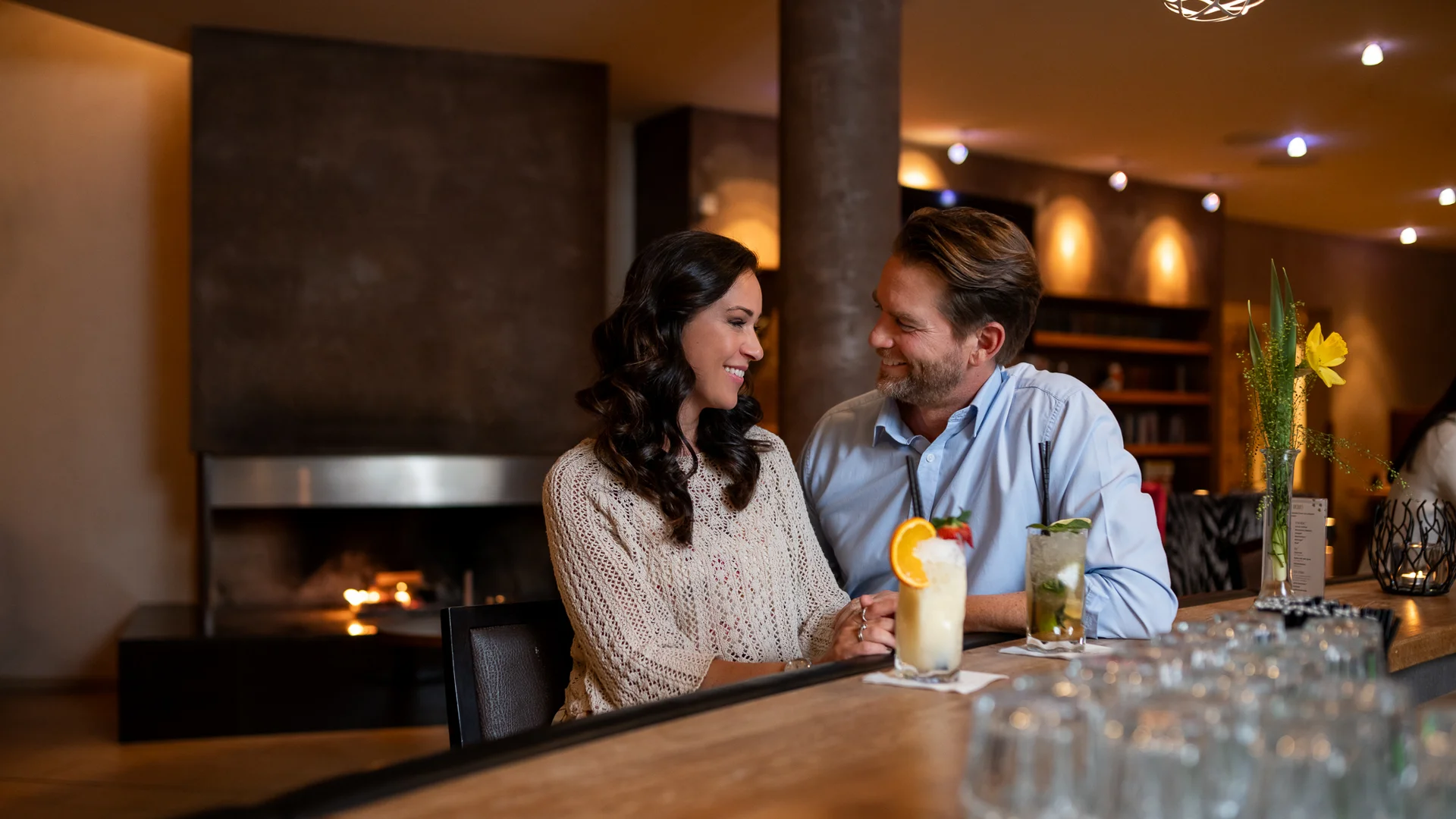 Paar sitzt an Bar mit Cocktails in gemütlicher Bar mit Kamin im Hintergrund