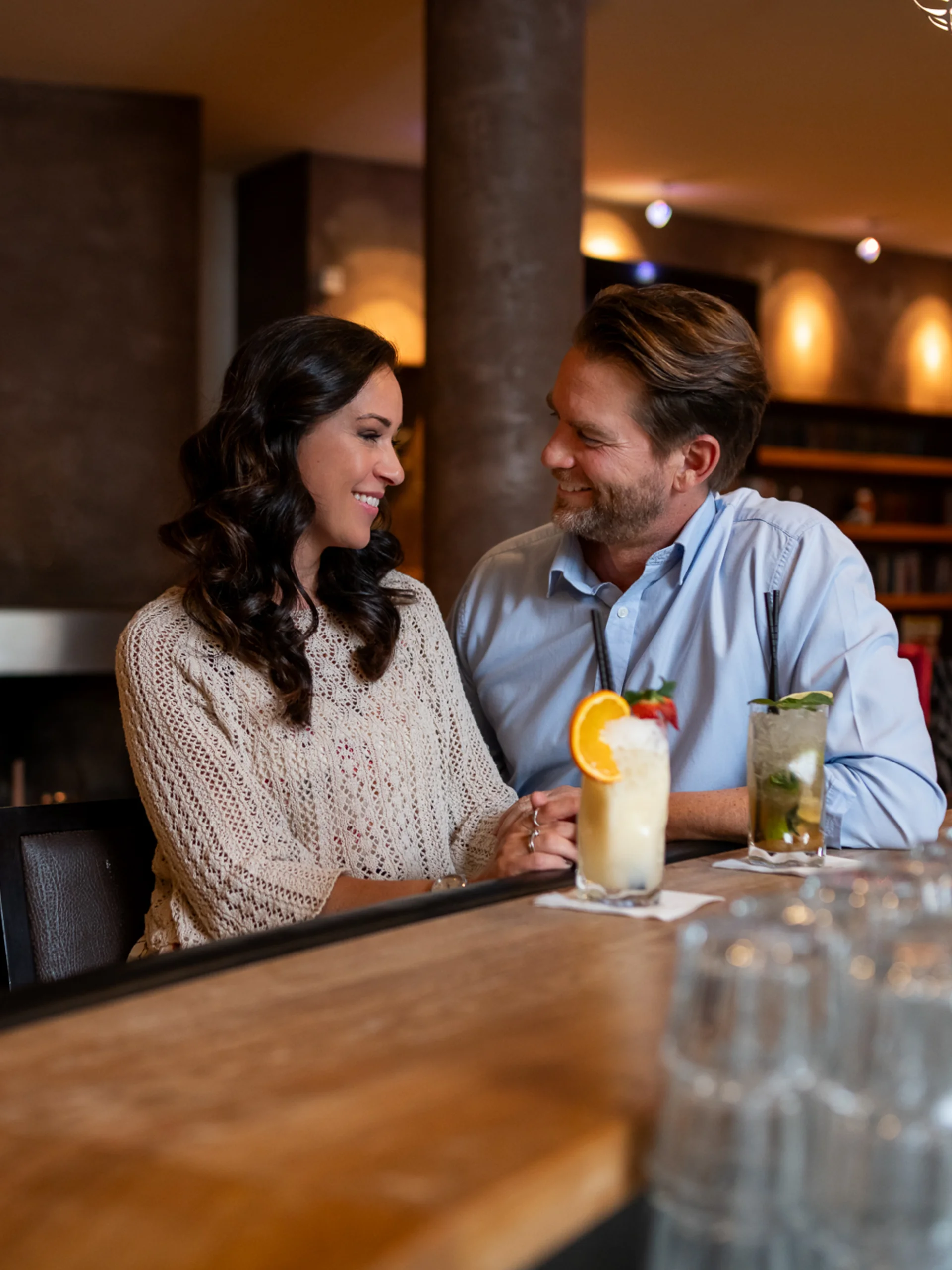 Paar sitzt an Bar mit Cocktails in gemütlicher Bar mit Kamin im Hintergrund
