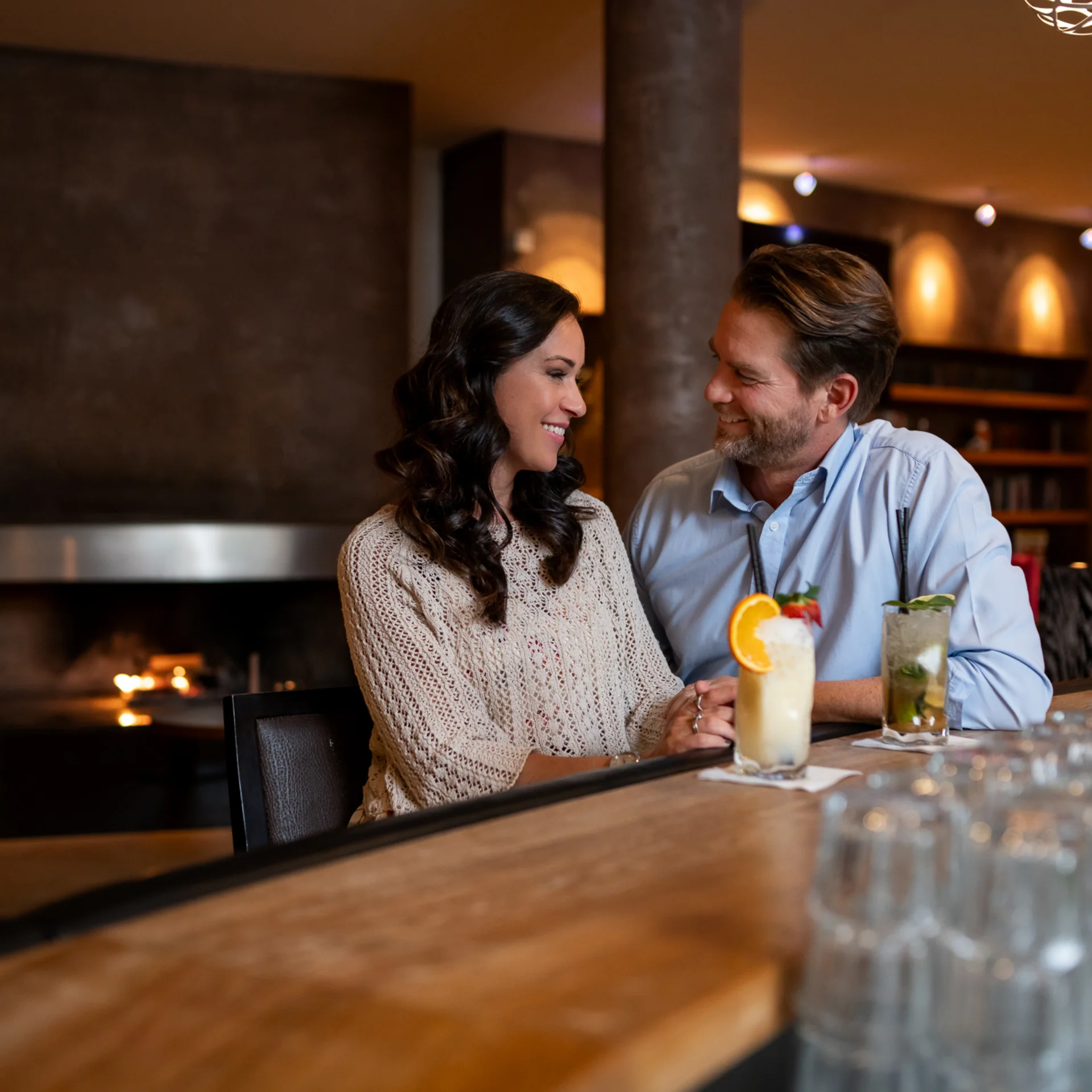 Paar sitzt an Bar mit Cocktails in gemütlicher Bar mit Kamin im Hintergrund