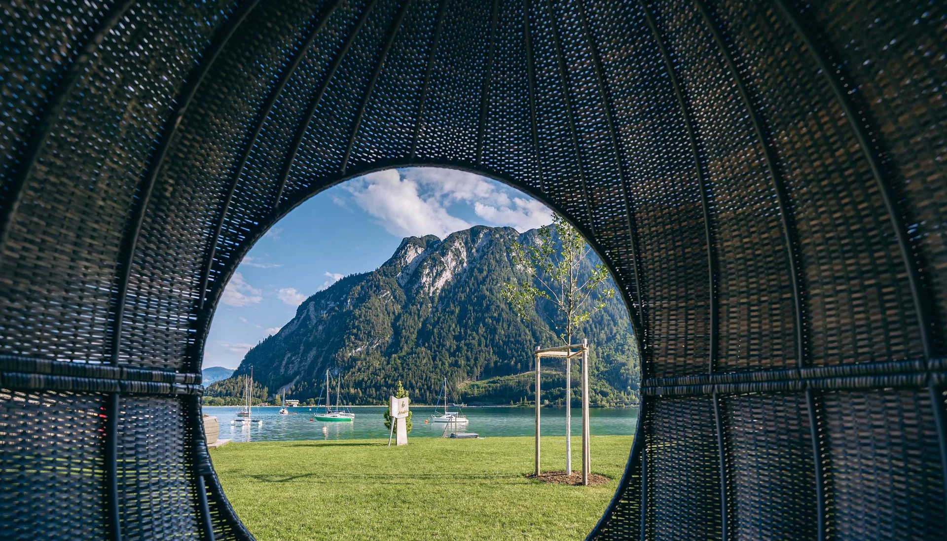 Blick durch ein rundes Korbgewebe auf Berg, See und Wiese mit Segelbooten