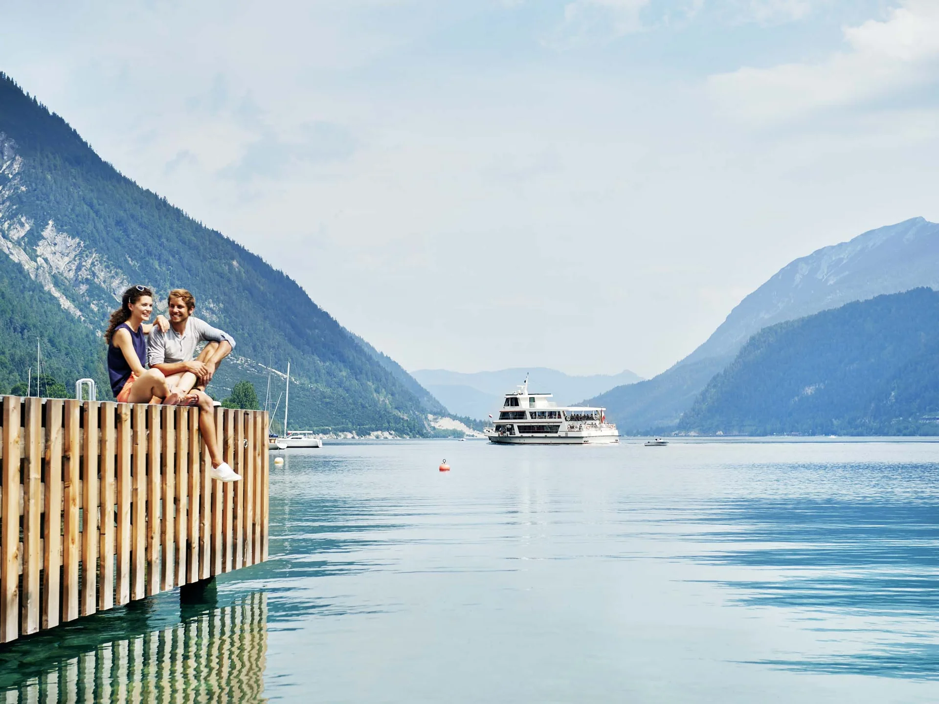 Paar sitzt am Steg eines Sees mit Berglandschaft und Ausflugsboot im Hintergrund