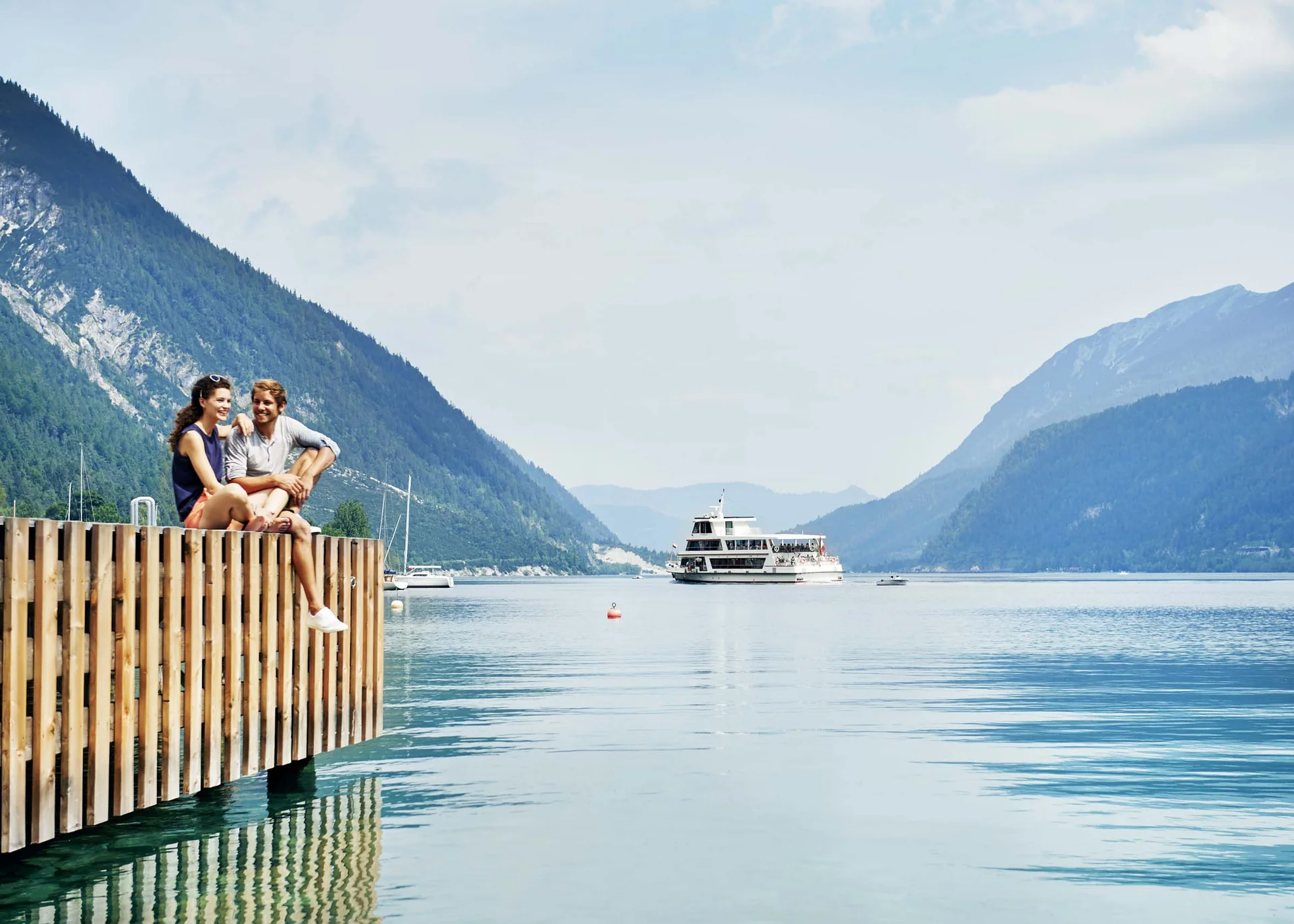 Paar sitzt am Steg eines Sees mit Berglandschaft und Ausflugsboot im Hintergrund