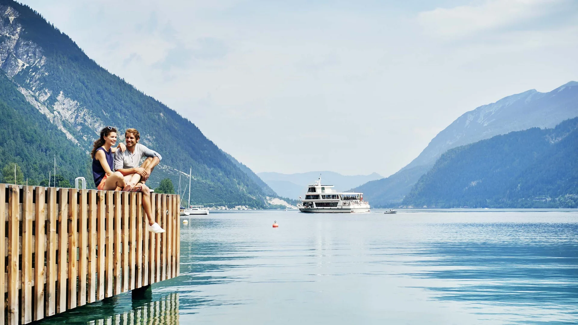 Paar sitzt am Steg eines Sees mit Berglandschaft und Ausflugsboot im Hintergrund
