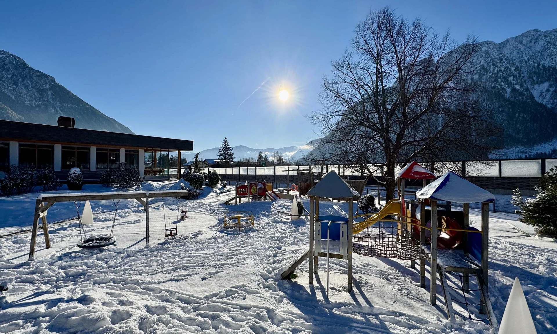 Schneebedeckter Kinderspielplatz mit Rutschen, Schaukel und Bergen im Hintergrund