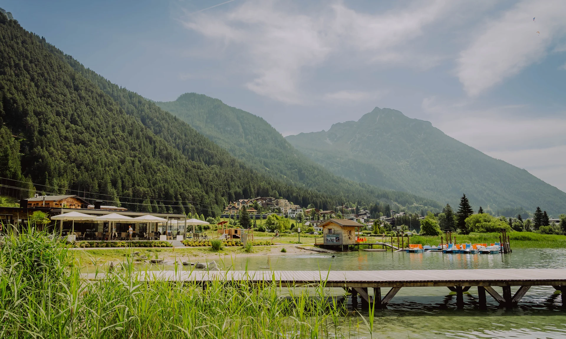 Steg an einem Bergsee mit Booten, Häusern und bewaldeten Bergen im Hintergrund