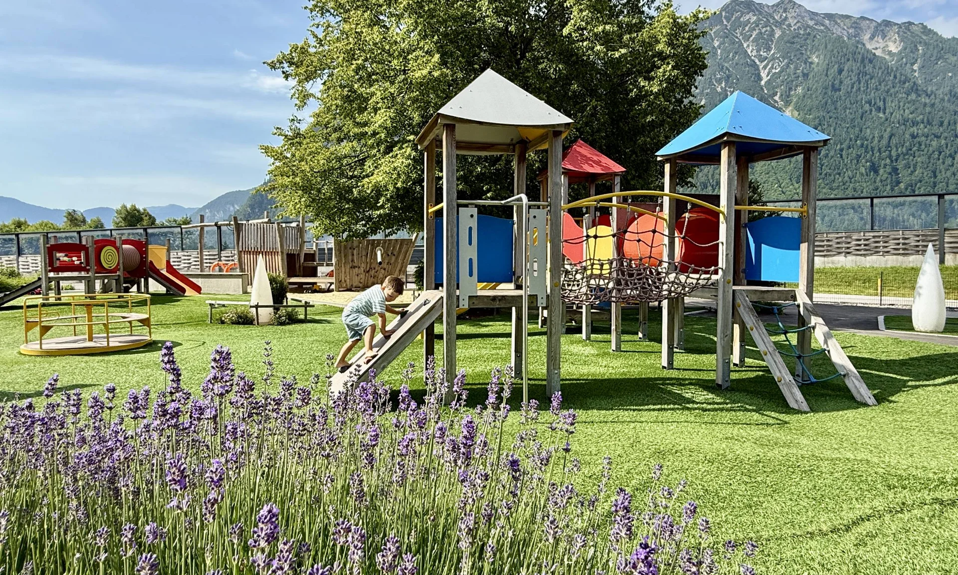 Kind klettert auf Spielplatz mit bunten Geräten vor Bergkulisse