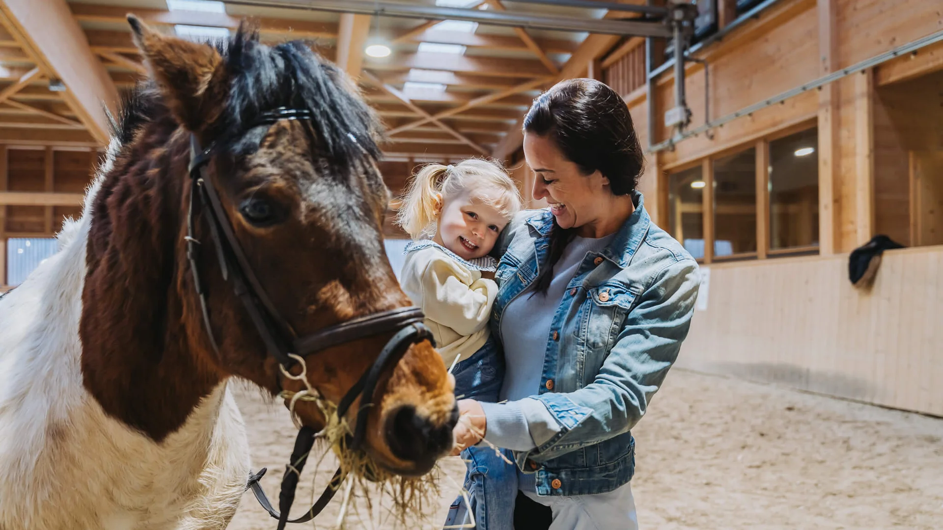 Frau hält lachendes Mädchen im Innenstall neben fressendem Pony