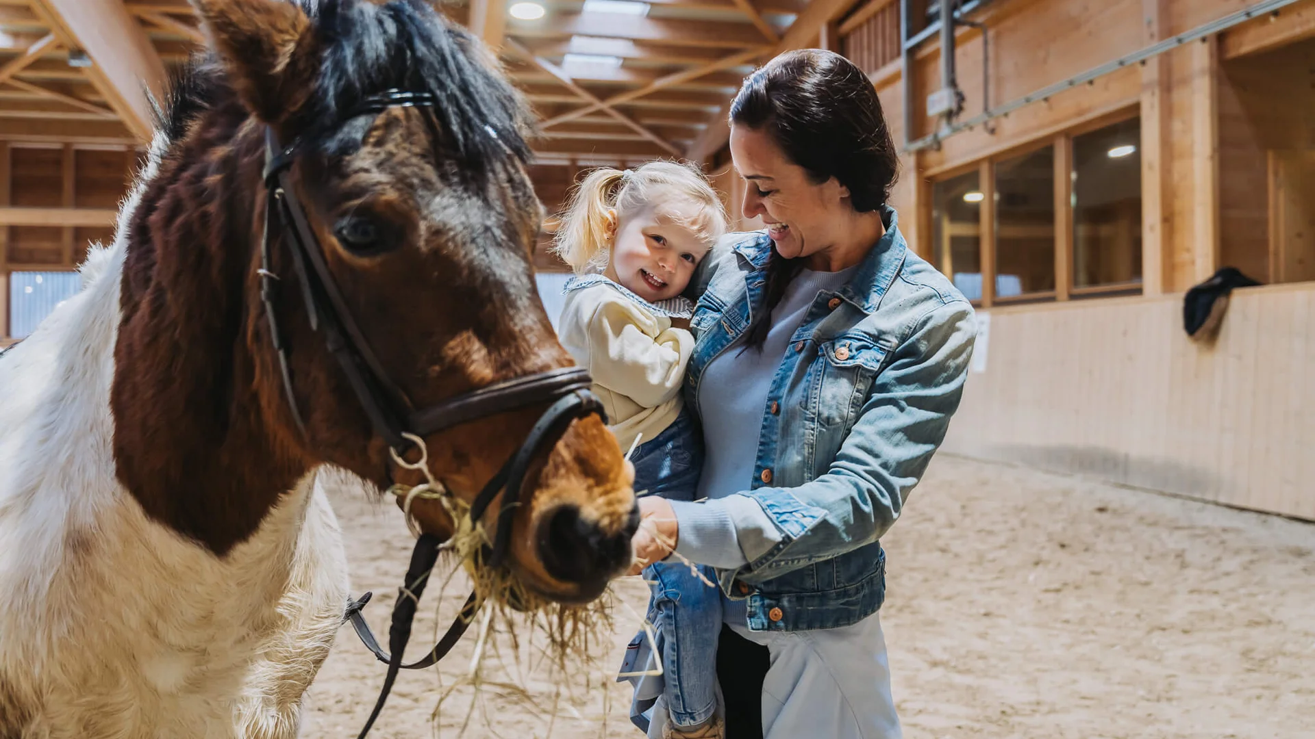 The family resort in Buchau for quality time in Tyrol Woman holding smiling girl in indoor stable next to pony eating hay