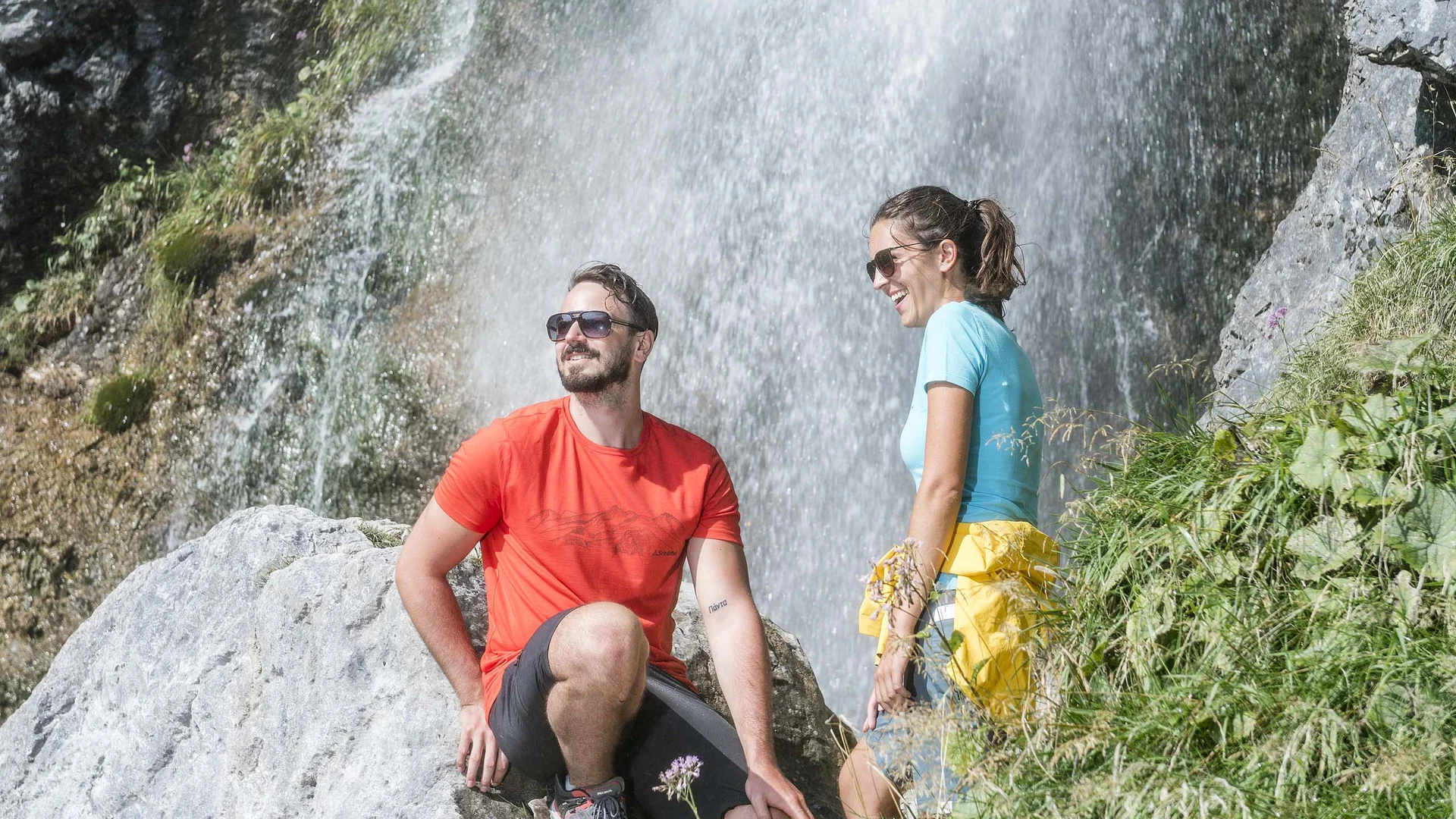 Mann in rotem Shirt und Frau in blauem Shirt sitzen neben Wasserfall auf Felsen