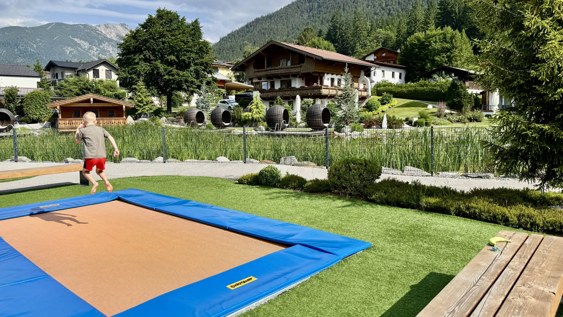 Kind springt auf Trampolin in bergigem Garten vor traditionellen Häusern