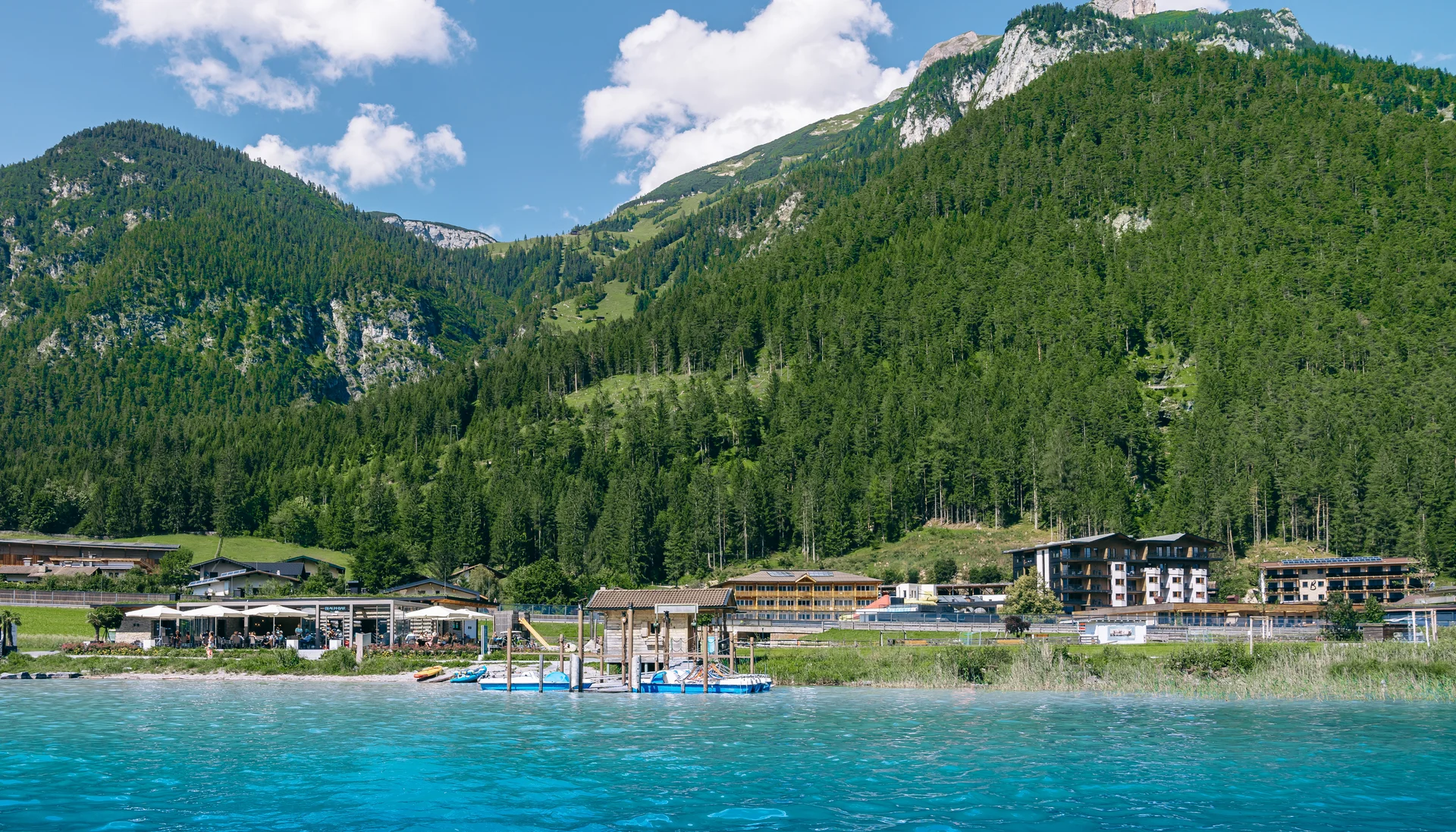 Berge und Wald hinter einem See mit Häusern und Steg am Ufer unter blauem Himmel