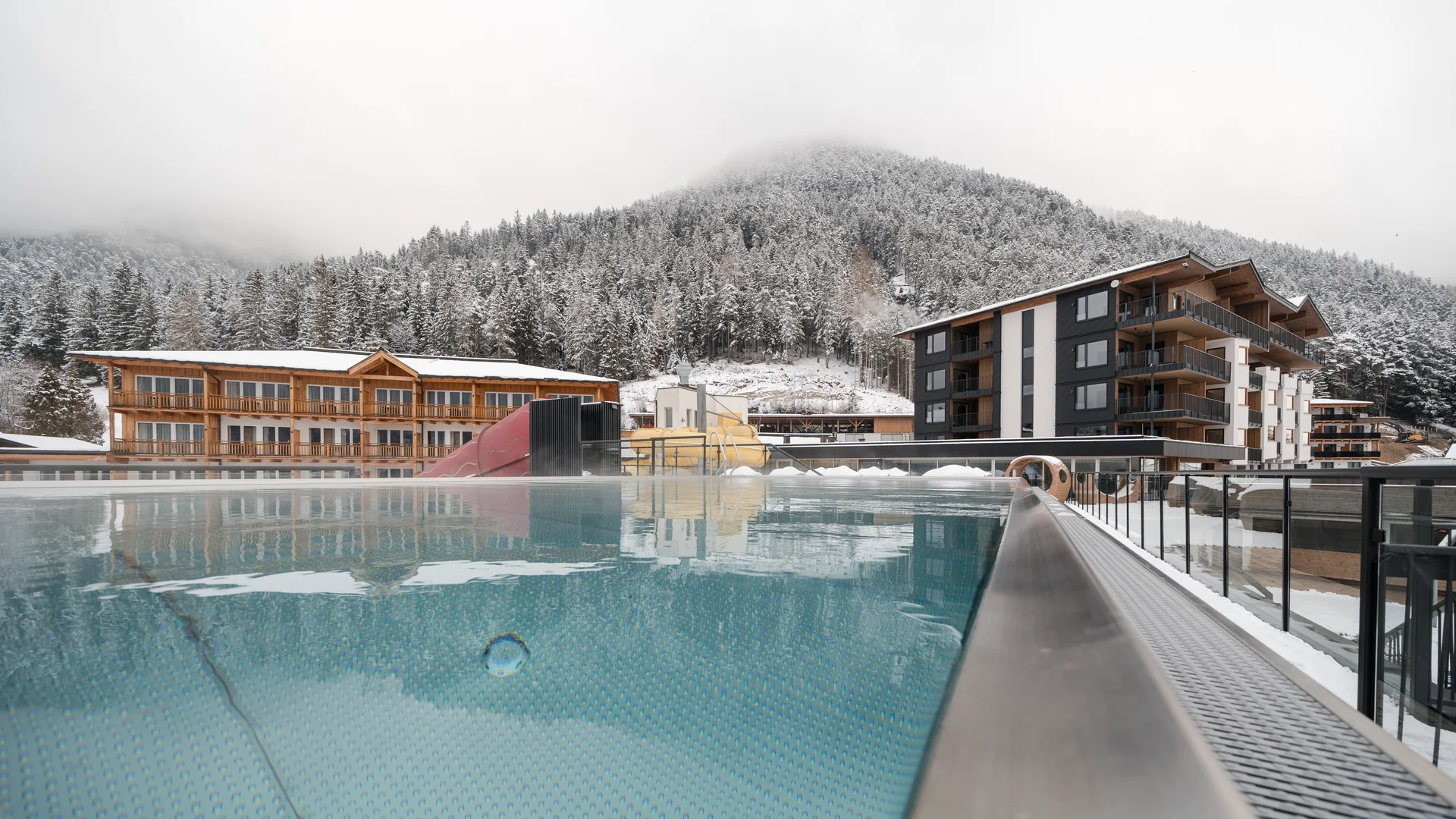 Outdoor pool with slide in front of snowy mountains and modern hotels