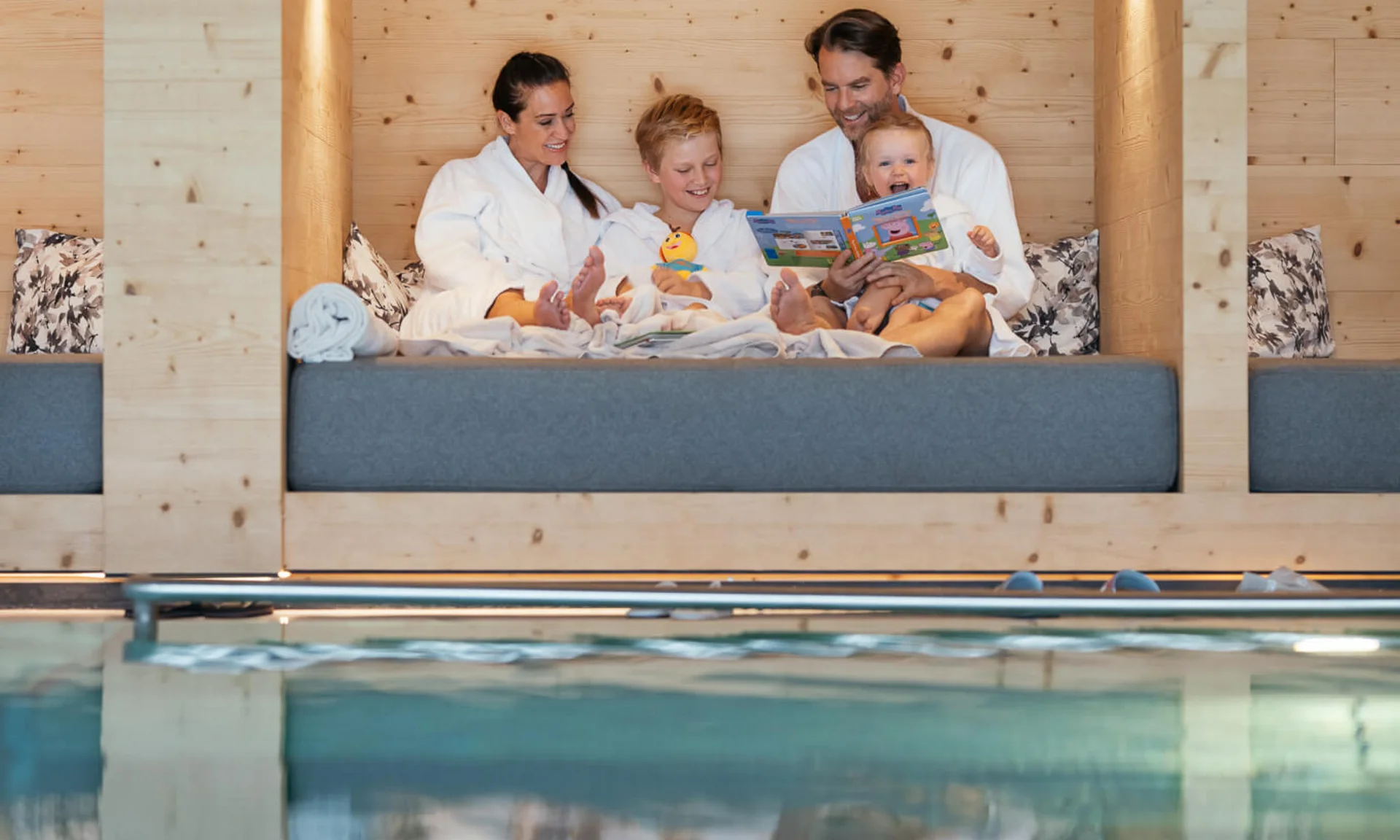 Familie in Bademänteln sitzt gemütlich an einem Holzruheraum neben dem Pool